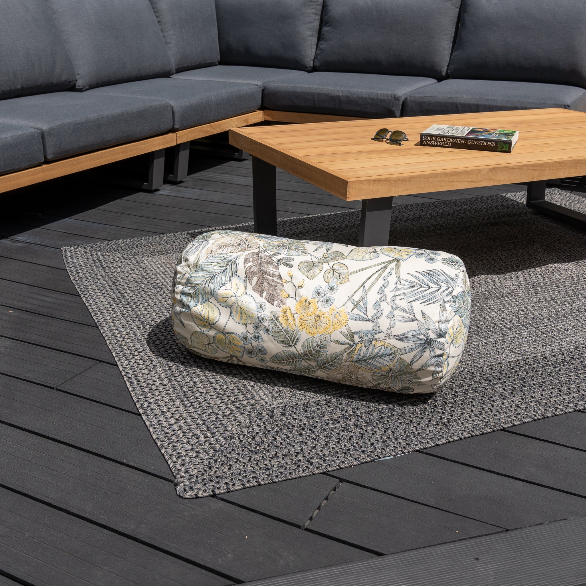 An Acrisol Bouquet Neutrals Large Round Bolster Floor Cushion (72cm x 35cm) is on a patio rug, complemented by solution-dyed acrylic outdoor scatter cushions, with a wooden table and gray outdoor sectional in the background.