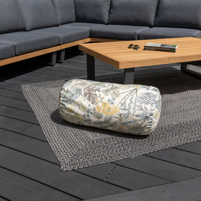 An Acrisol Bouquet Neutrals Large Round Bolster Floor Cushion (72cm x 35cm) is on a patio rug, complemented by solution-dyed acrylic outdoor scatter cushions, with a wooden table and gray outdoor sectional in the background.