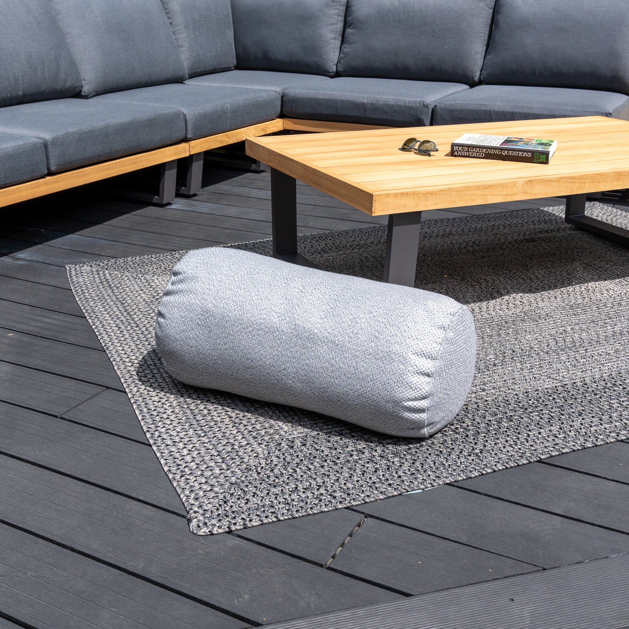 An Agora Senda Ceniza Large Round Bolster Floor Cushion – 72cm x 35cm, rests on a woven rug next to a wooden coffee table and a gray sectional couch on the deck, as part of a set of outdoor scatter cushions. Enhance longevity with the Golden Care Fabrics & Upholstery Care Kit.