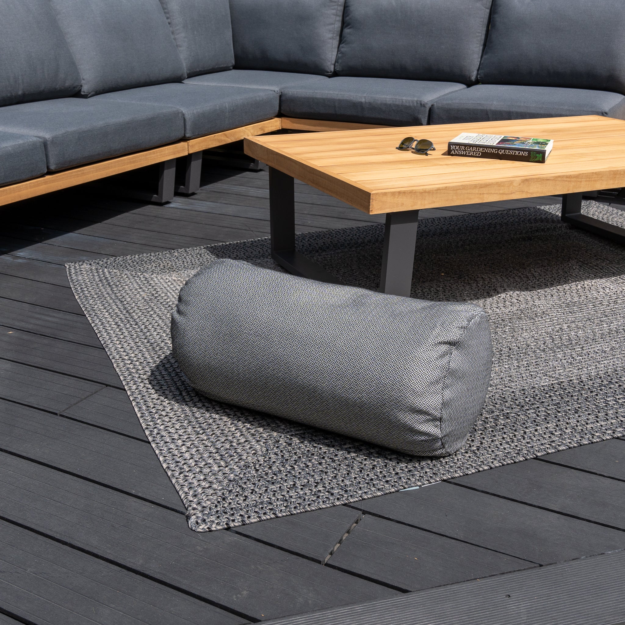 Outdoor seating area with grey, solution-dyed acrylic cushions, a wooden coffee table, a book, sunglasses, and an Agora Vimini Pirita Large Round Bolster Floor Cushion - 72cm x 35cm on a rug.