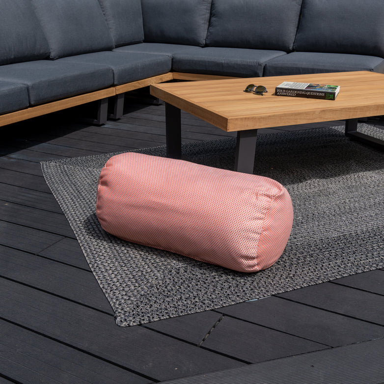 An Agora Scala Coral Large Round Bolster Floor Cushion (72cm x 35cm) on a gray rug in front of a wooden coffee table and a gray sectional outdoor sofa, complemented by vibrant outdoor scatter cushions.