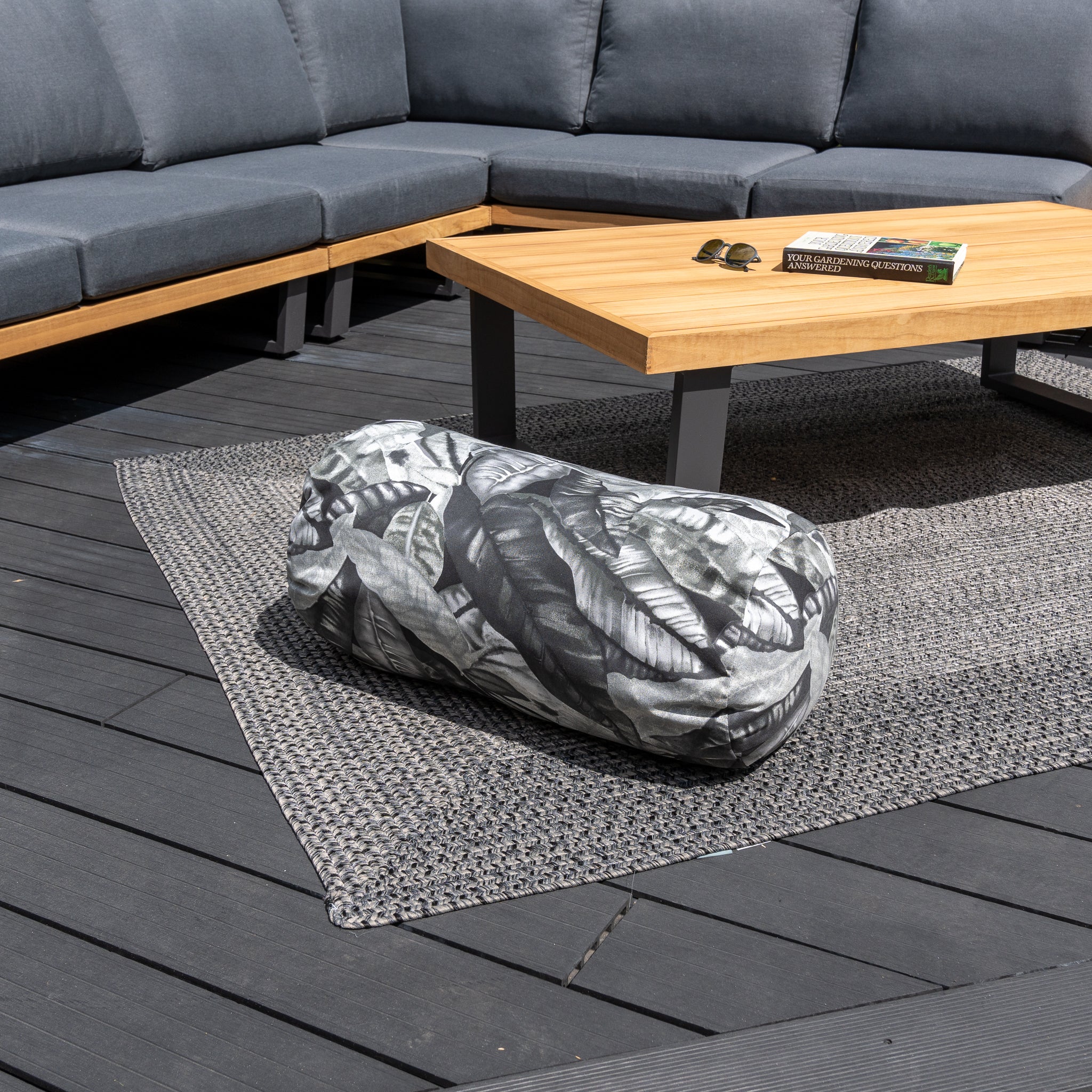 Outdoor furniture set featuring a wooden table, the Acrisol Amazonia Gris Large Round Bolster Floor Cushion - 72cm x 35cm, a book on a woven rug, and gray pieces arranged on the deck. Ideal for relaxation, this setup ensures durability with solution-dyed acrylic while outdoor scatter cushions add comfort.