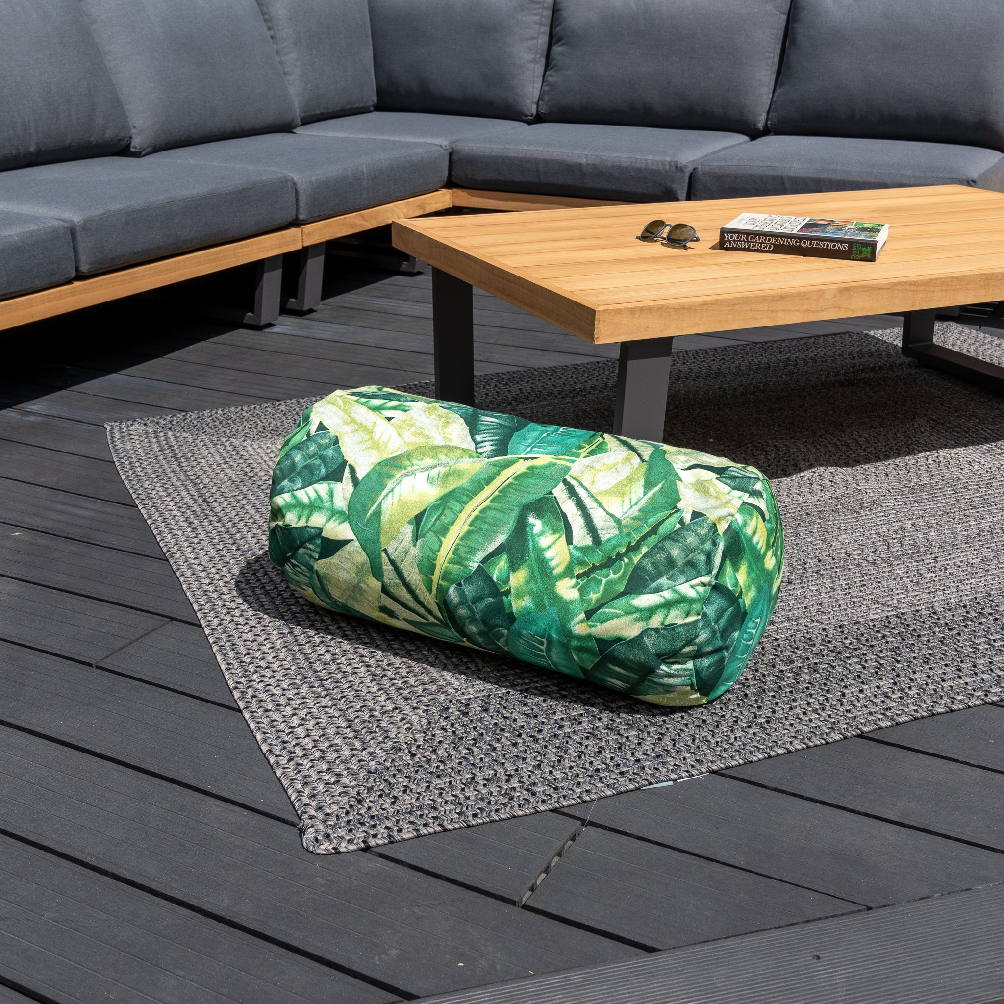 A patio with a gray sectional, outdoor scatter cushions made from the highest quality solution-dyed acrylic, an outdoor rug, a wooden table, and an Acrisol Amazonia Verde Large Round Bolster Floor Cushion featuring a green leaf pattern.