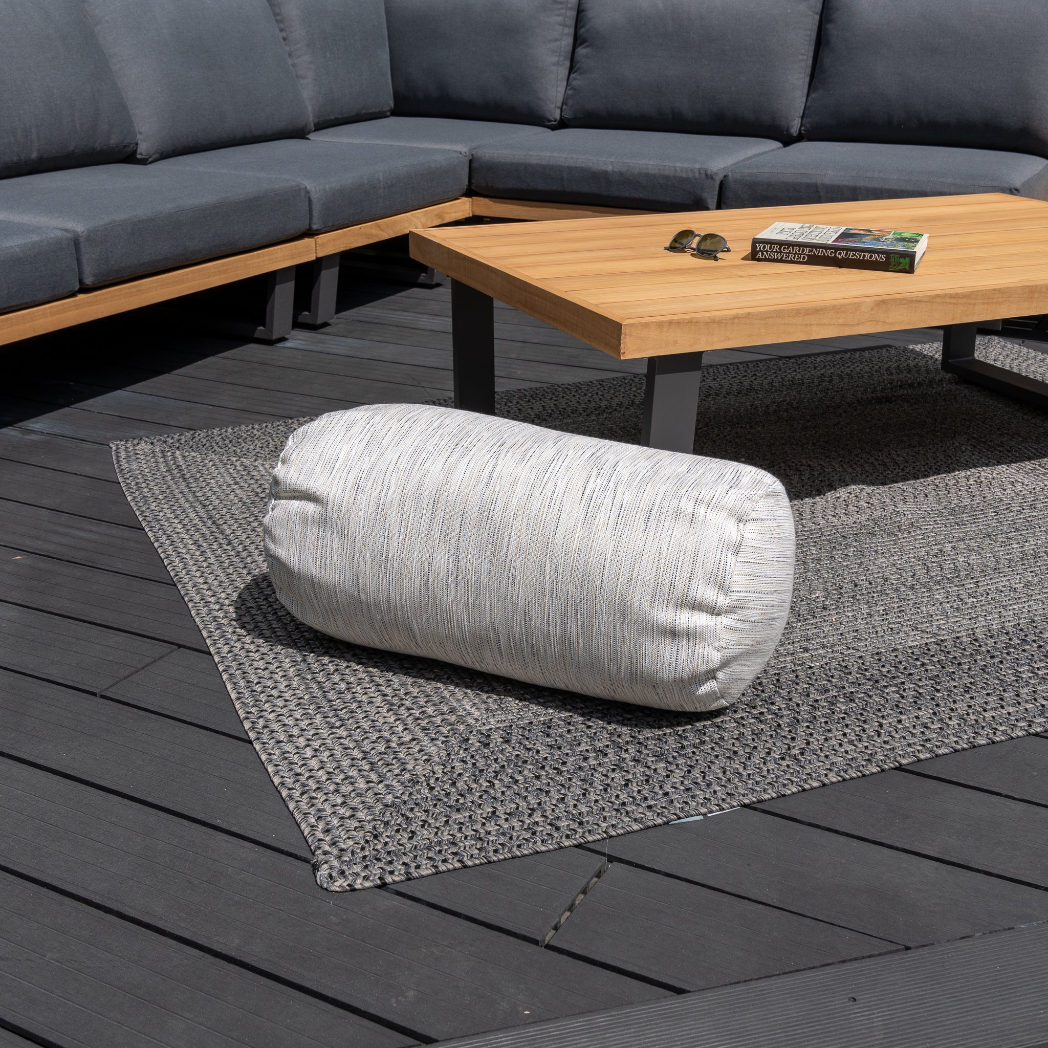 An Agora Texture Ugo Large Round Bolster Floor Cushion (72cm x 35cm) on a textured rug beside a wooden table and cushioned outdoor seating enhances garden personalization with vibrant outdoor scatter cushions.