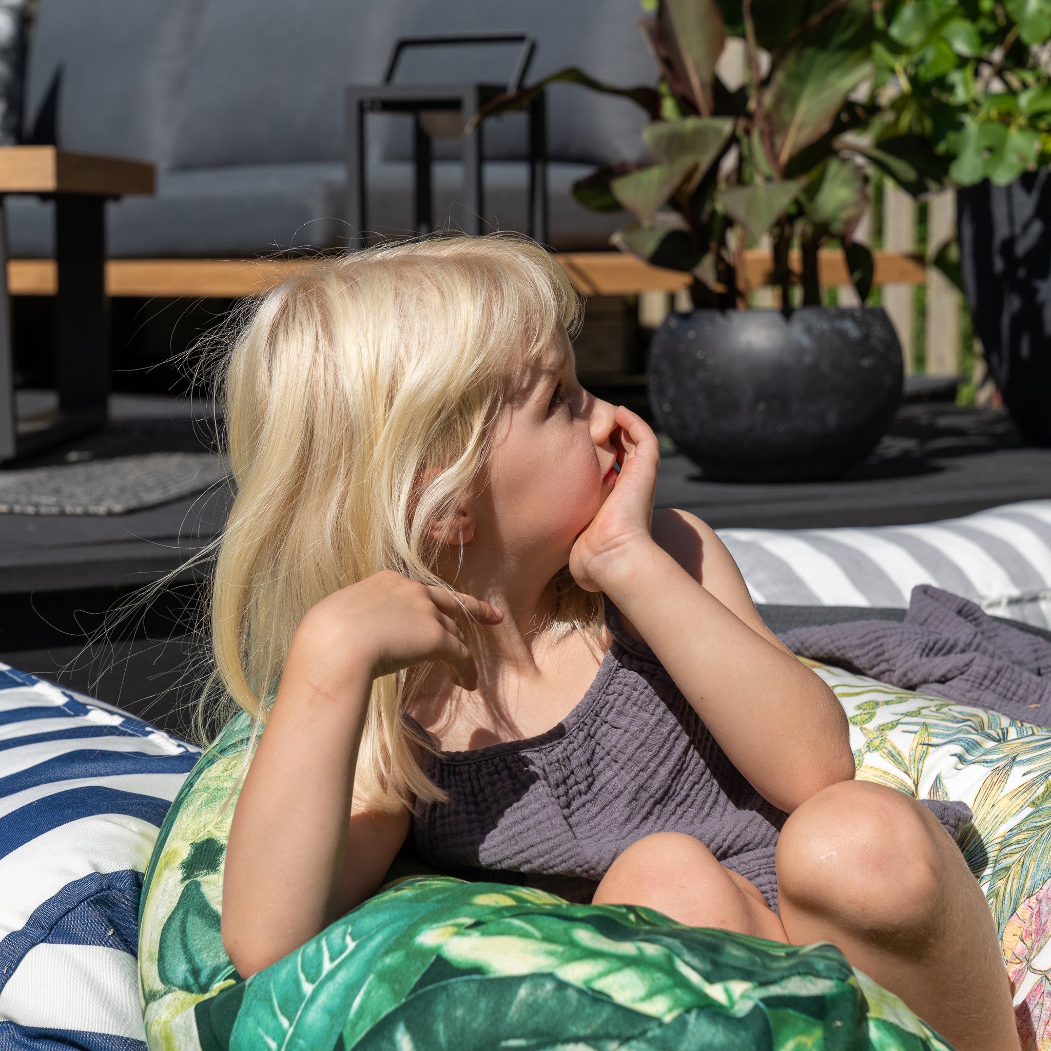 A young girl with blonde hair sits on an Acrisol Amazonia Verde Large Round Bolster Floor Cushion, resting her chin on her hand against an outdoor backdrop of lush greenery. Crafted from the highest quality solution-dyed acrylic, these cushions ensure vibrant longevity in any weather.