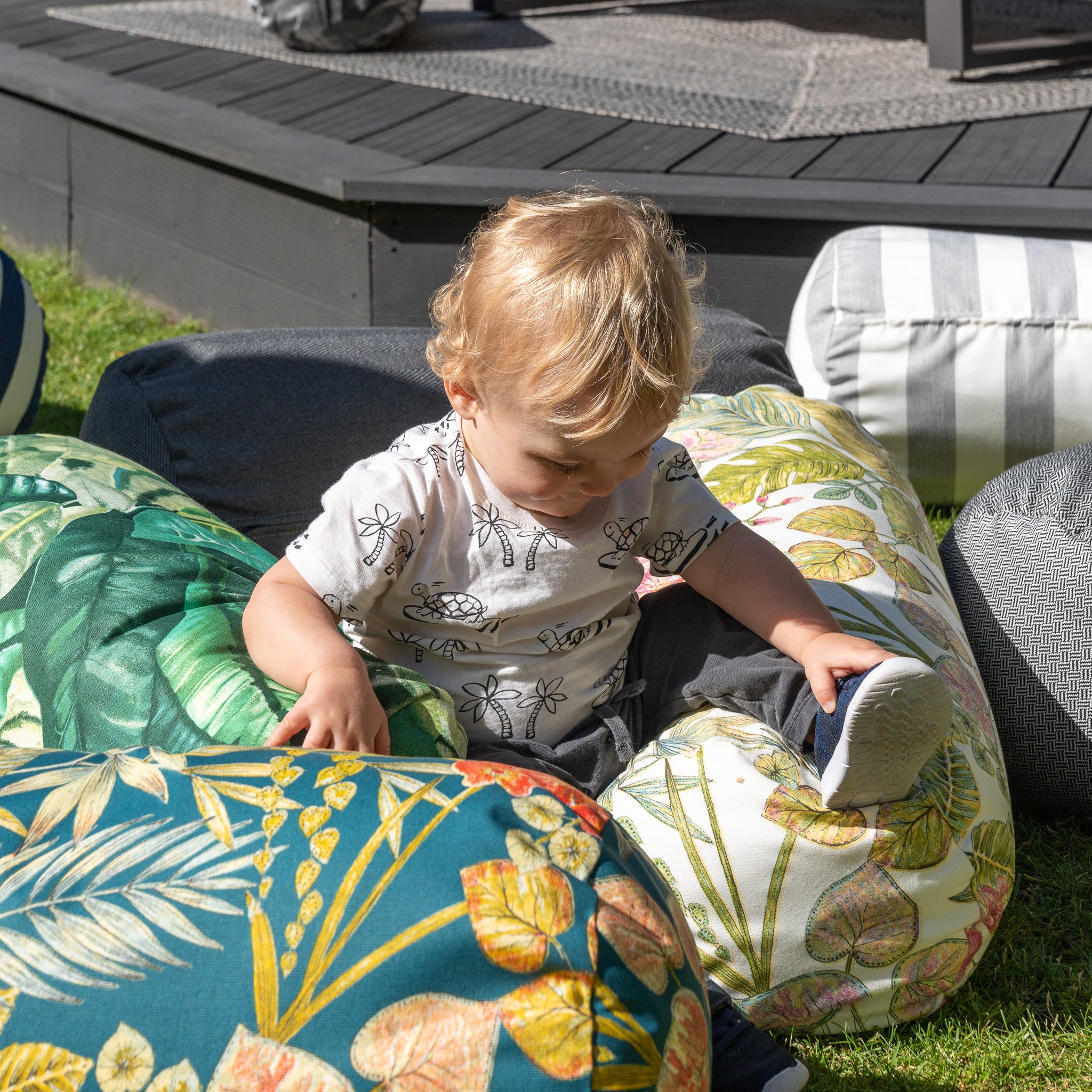 A toddler sits among colorful, leaf-patterned Acrisol Amazonia Verde Large Round Bolster Floor Cushions made from the highest quality solution-dyed acrylic on a grassy lawn, looking down and touching the cushions.