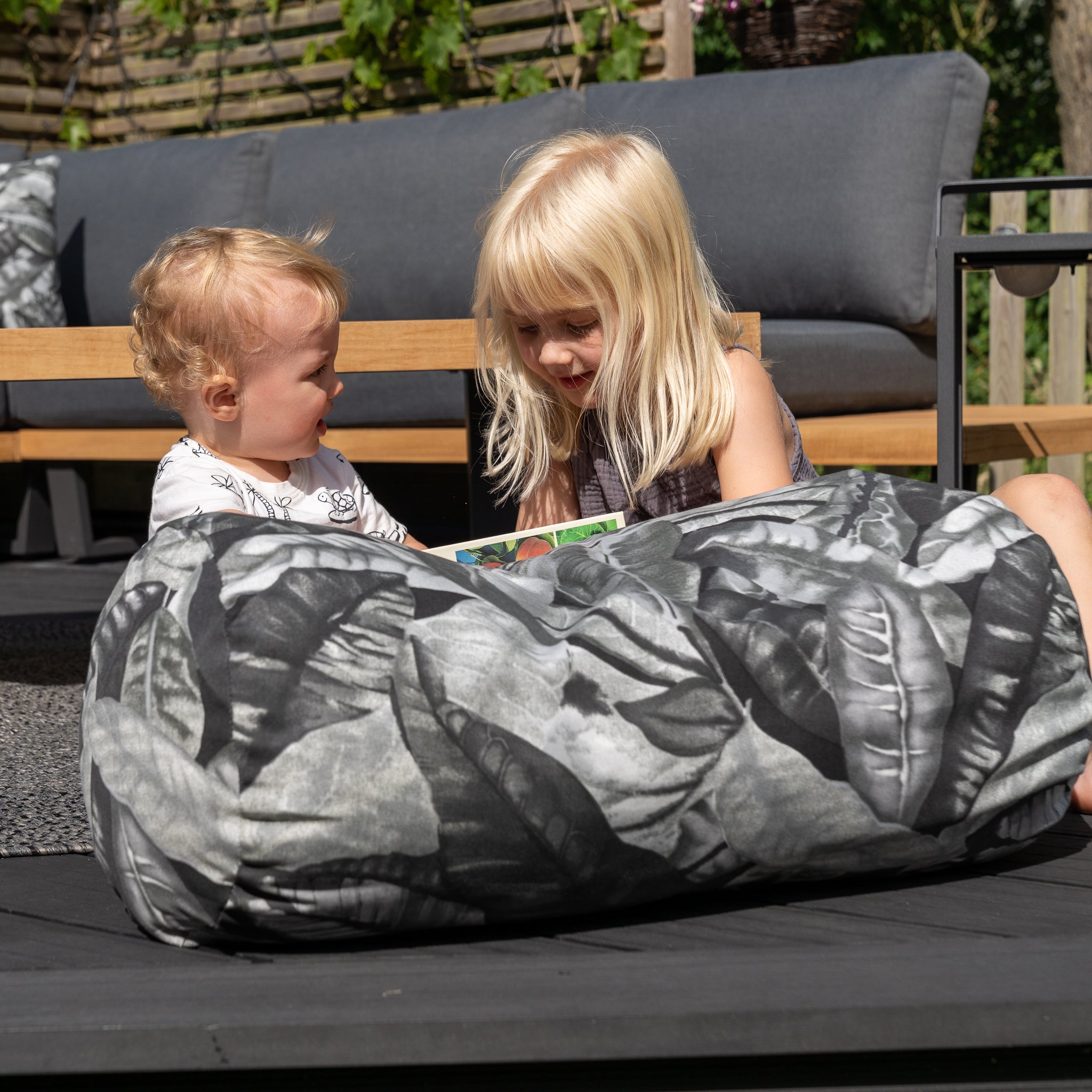 Two children sitting on a patio, with one reading a book to the other while they both relax on an Acrisol Amazonia Gris Large Round Bolster Floor Cushion - 72cm x 35cm from their outdoor furniture set.