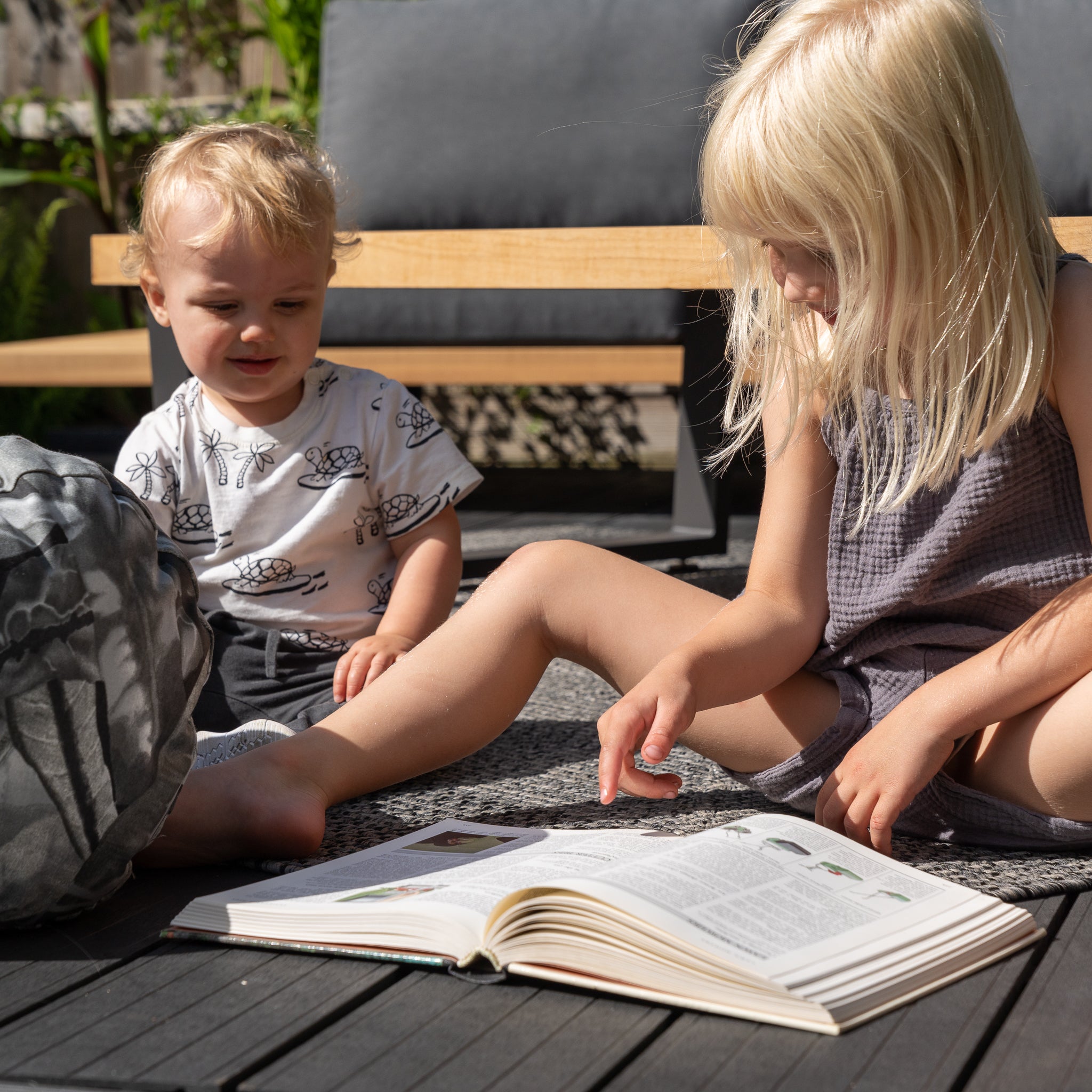 Two young children sit outdoors on an Acrisol Amazonia Gris Large Round Bolster Floor Cushion - 72cm x 35cm, with one reading a book to the other. They are comfortably seated on wooden deck flooring in sunlight, surrounded by vibrant outdoor scatter cushions.