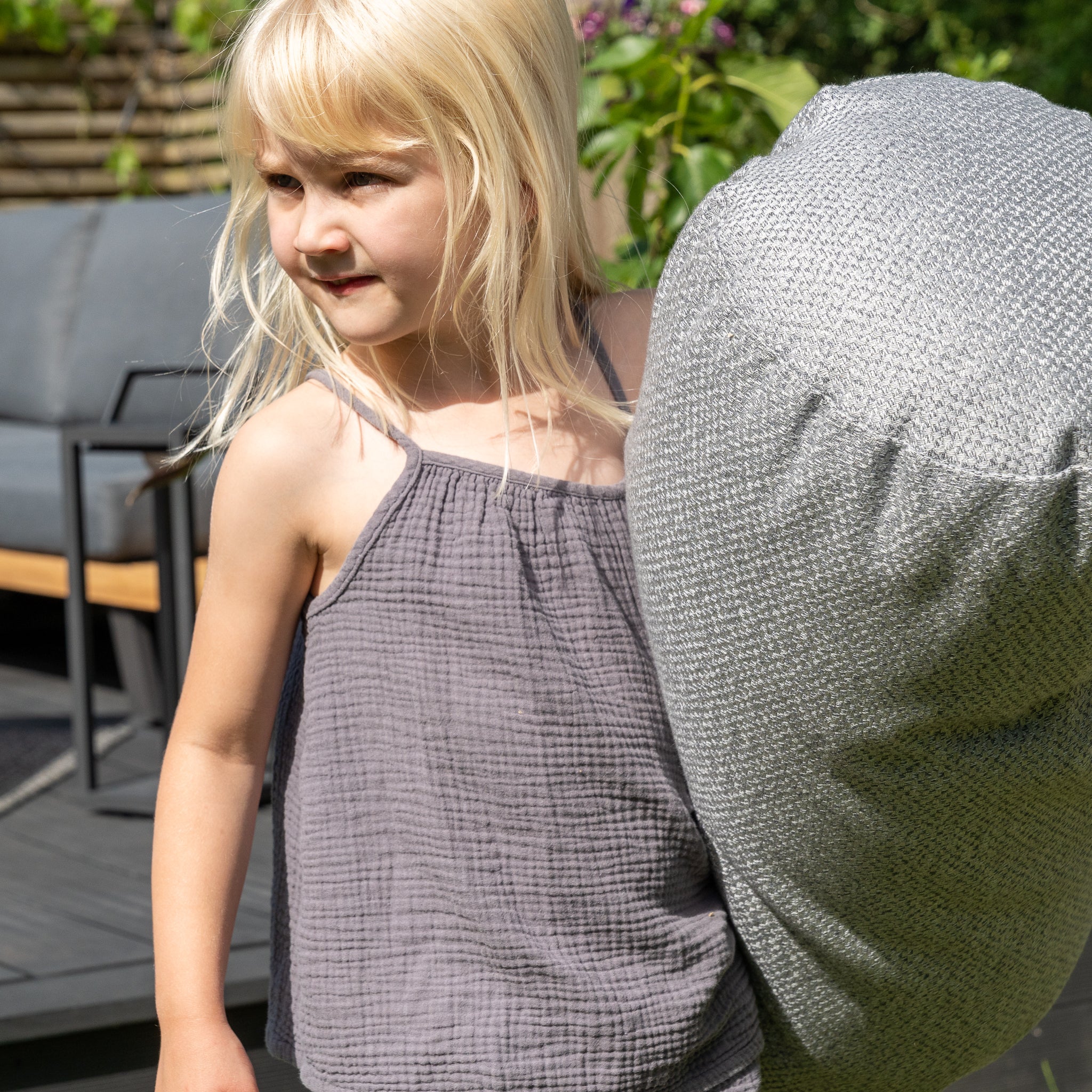 A young girl with blonde hair, dressed in a grey outfit, holds an Agora Senda Ceniza Large Round Bolster Floor Cushion - 72cm x 35cm outdoors, relishing the comfort of her outdoor scatter cushion.