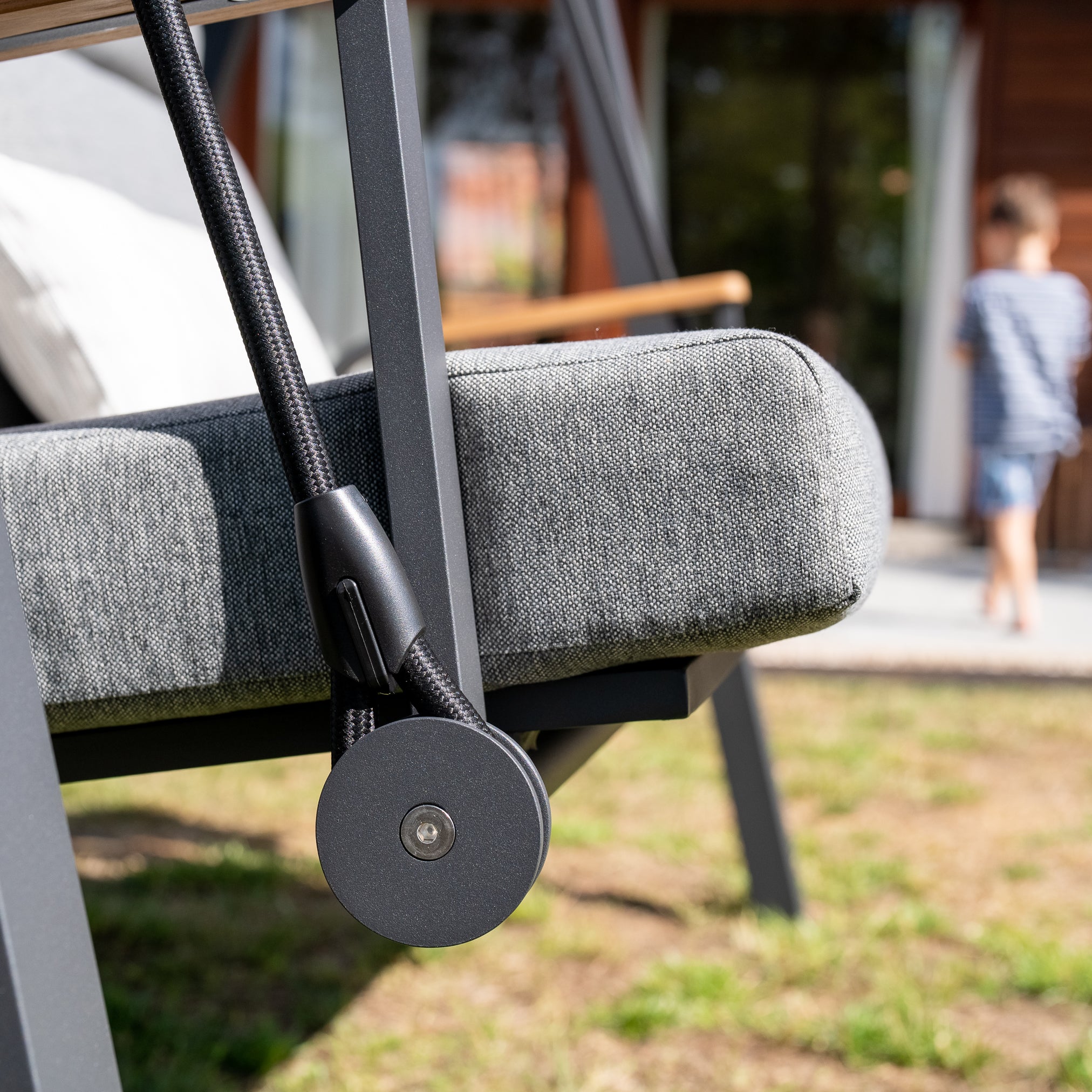 Close-up of a washed gray Lima Deluxe Aluminium Swing Seat with intricate rope detail, expertly crafted from Olefin material; blurry background with a person walking away.