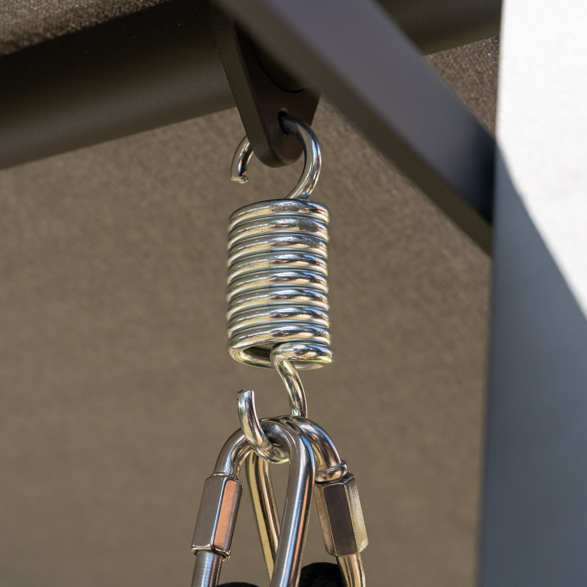 Close-up of a metal spring connected to hooks and a carabiner hanging from a metal frame, part of the robust design seen in the Lima Deluxe Aluminium Swing Seat in Washed Grey.