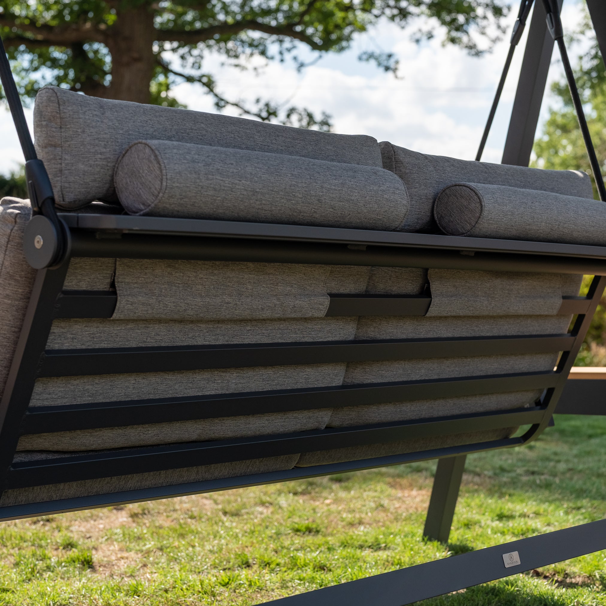 Close-up of a Lima Deluxe Aluminium Swing Seat in Washed Grey with a cushioned seat and a metal frame on a grassy area, surrounded by trees in the background.
