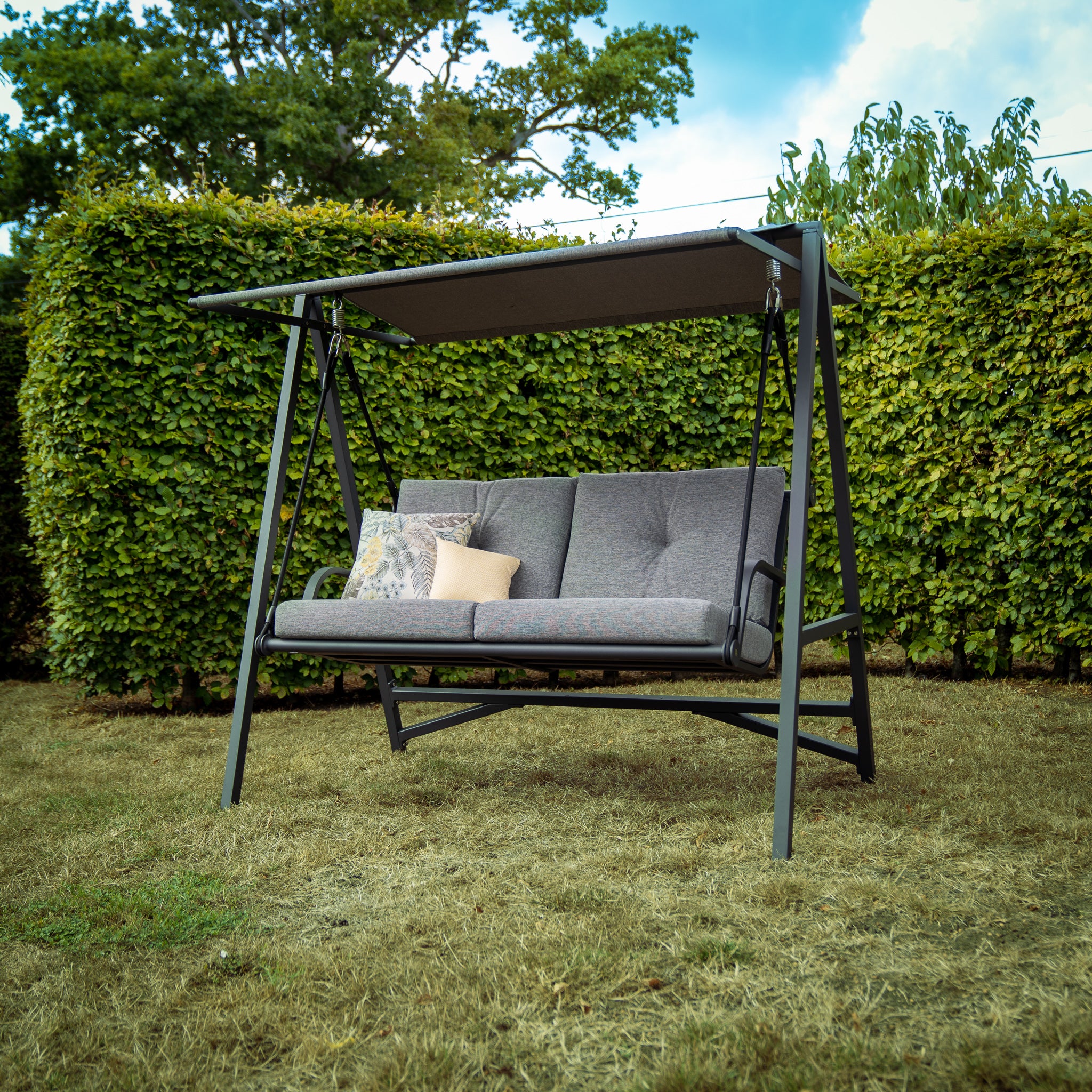 A cushioned Lima Aluminium Swing Seat in Washed Grey with an olefin canopy, placed on a lawn in front of a green hedge under a blue sky, features a durable powder-coated aluminium frame.