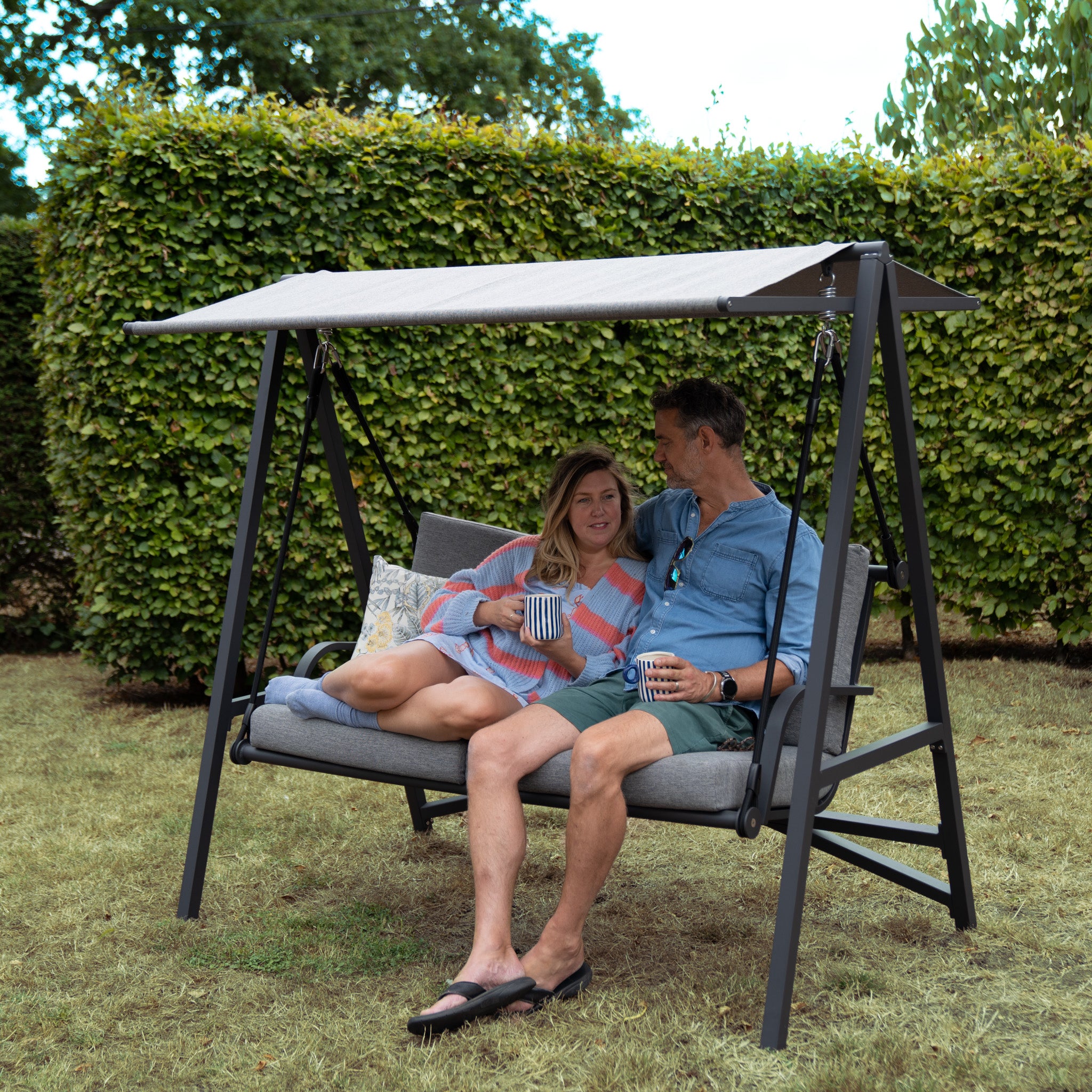 A couple sits on a Lima Aluminium Swing Seat in Washed Grey with an olefin canopy in a garden, smiling and holding mugs, surrounded by hedges and trees.