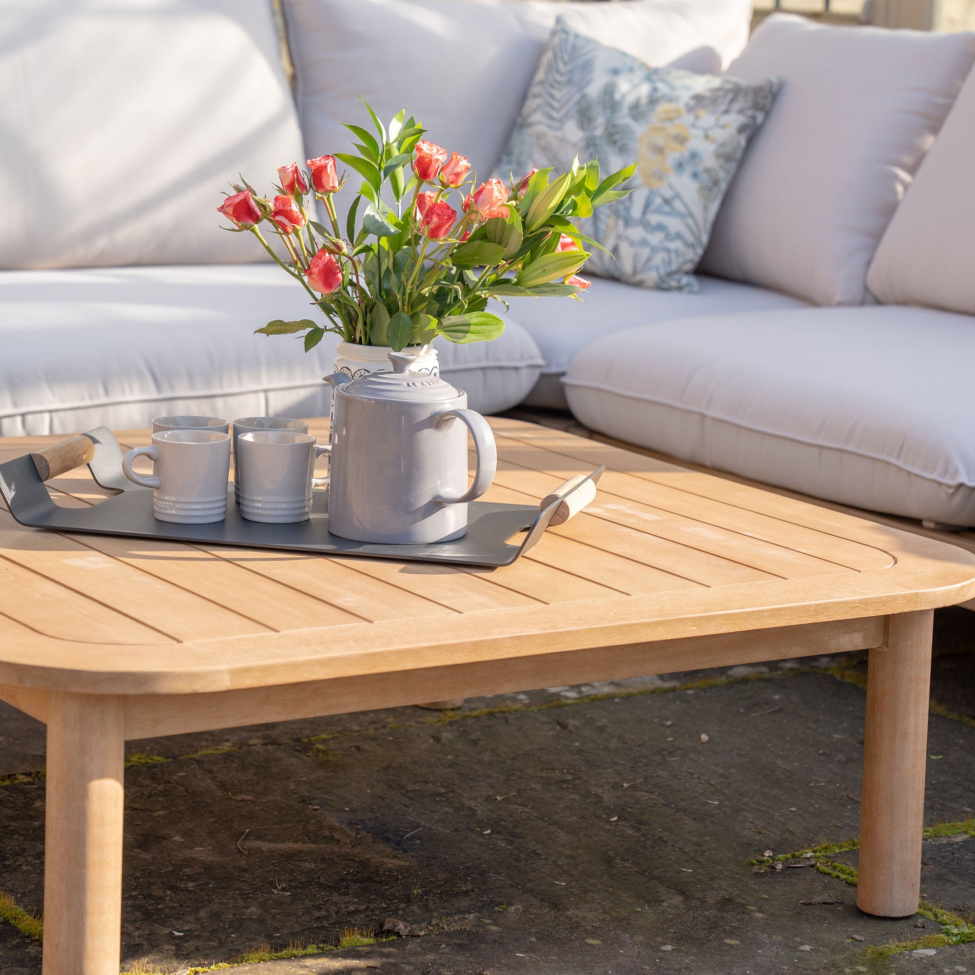 A Lavenham Corner Sofa Set in Linen from the Lavenham Collection is seen in the background, while a wooden coffee table crafted from sustainable eucalyptus wood holds a tray with a teapot, cups, and a vase of pink flowers.
