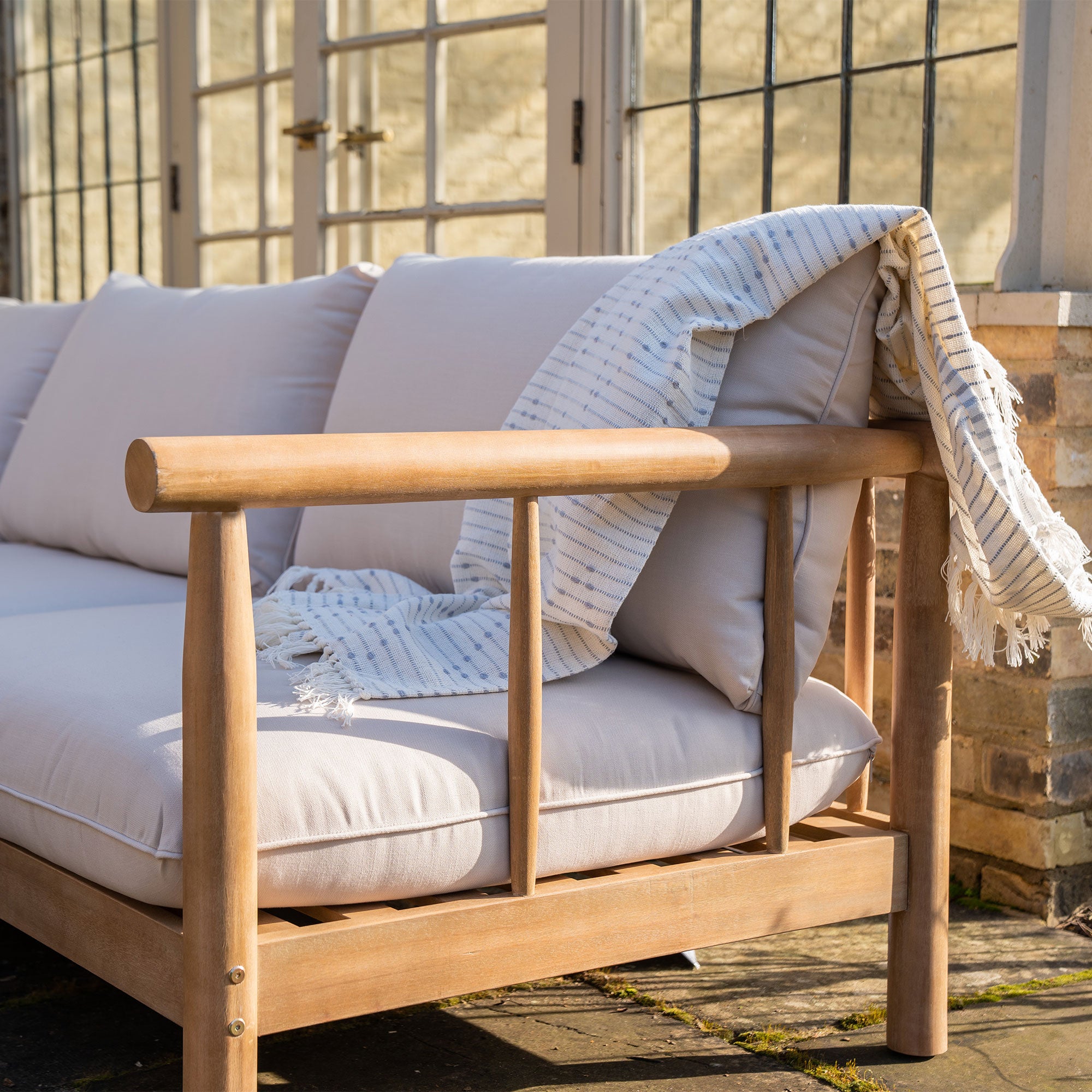 A Lavenham Corner Sofa Set in Linen, featuring light gray cushions and a striped throw blanket draped over the armrest, crafted from sustainable eucalyptus wood.