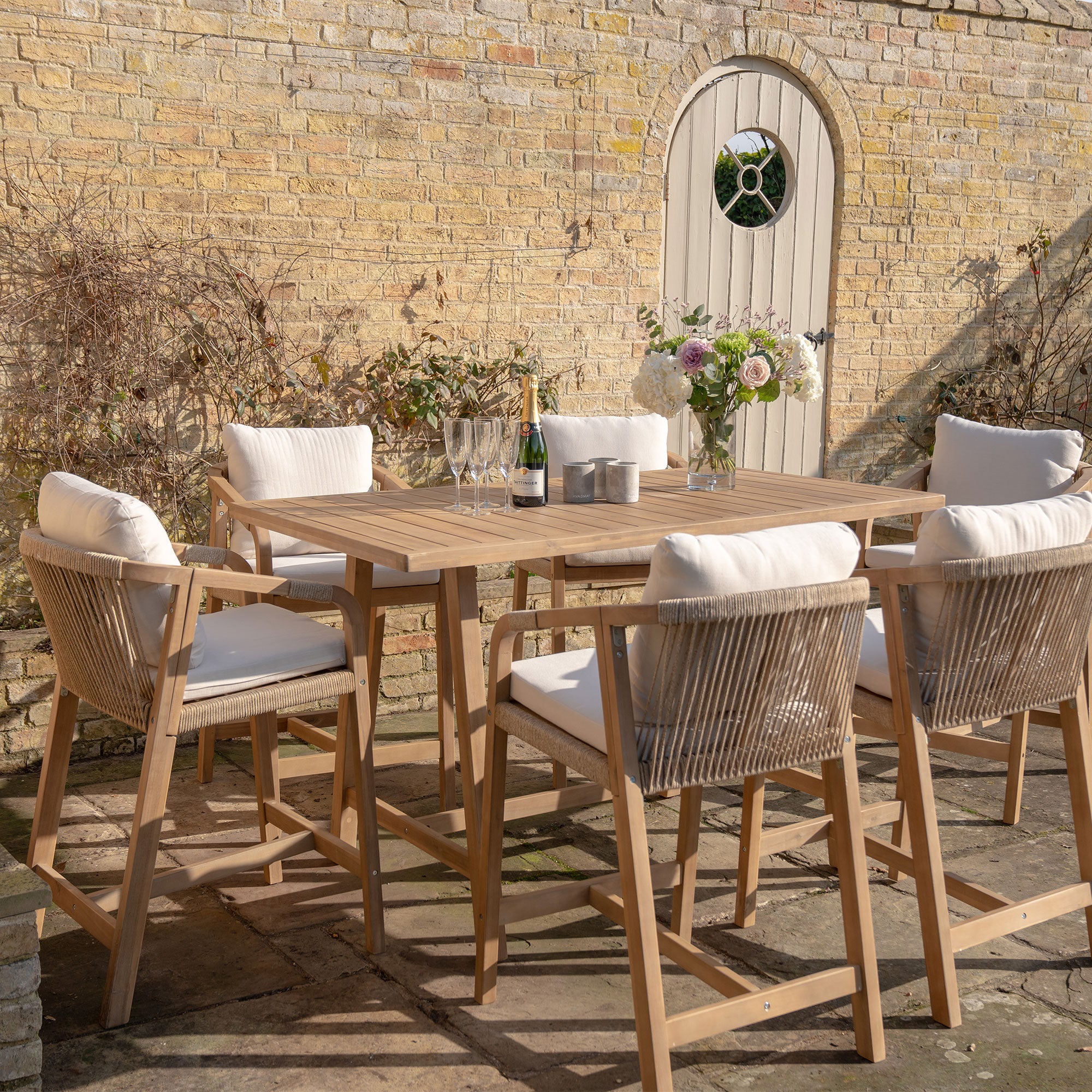 Outdoor patio featuring the Quay 6 Seat Rectangular Bar Set in Linen, made from sustainably sourced acacia wood, six cushioned chairs, flowers, and a bottle of wine in front of a brick wall.