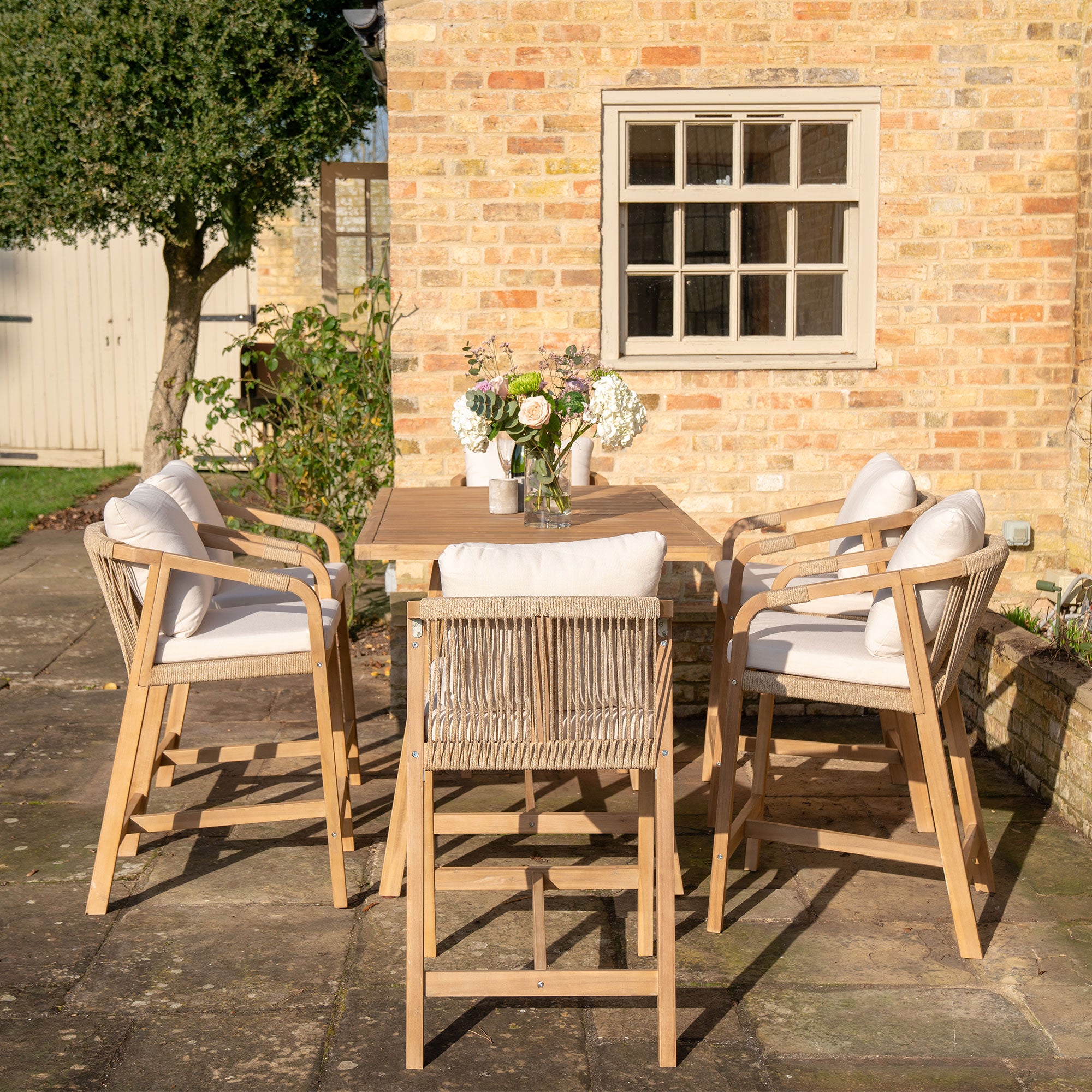 The Quay 6 Seat Rectangular Bar Set in Linen, featuring a sustainably sourced acacia wood table and cushioned chairs, is displayed against a brick wall with a tree and shed door in the background.