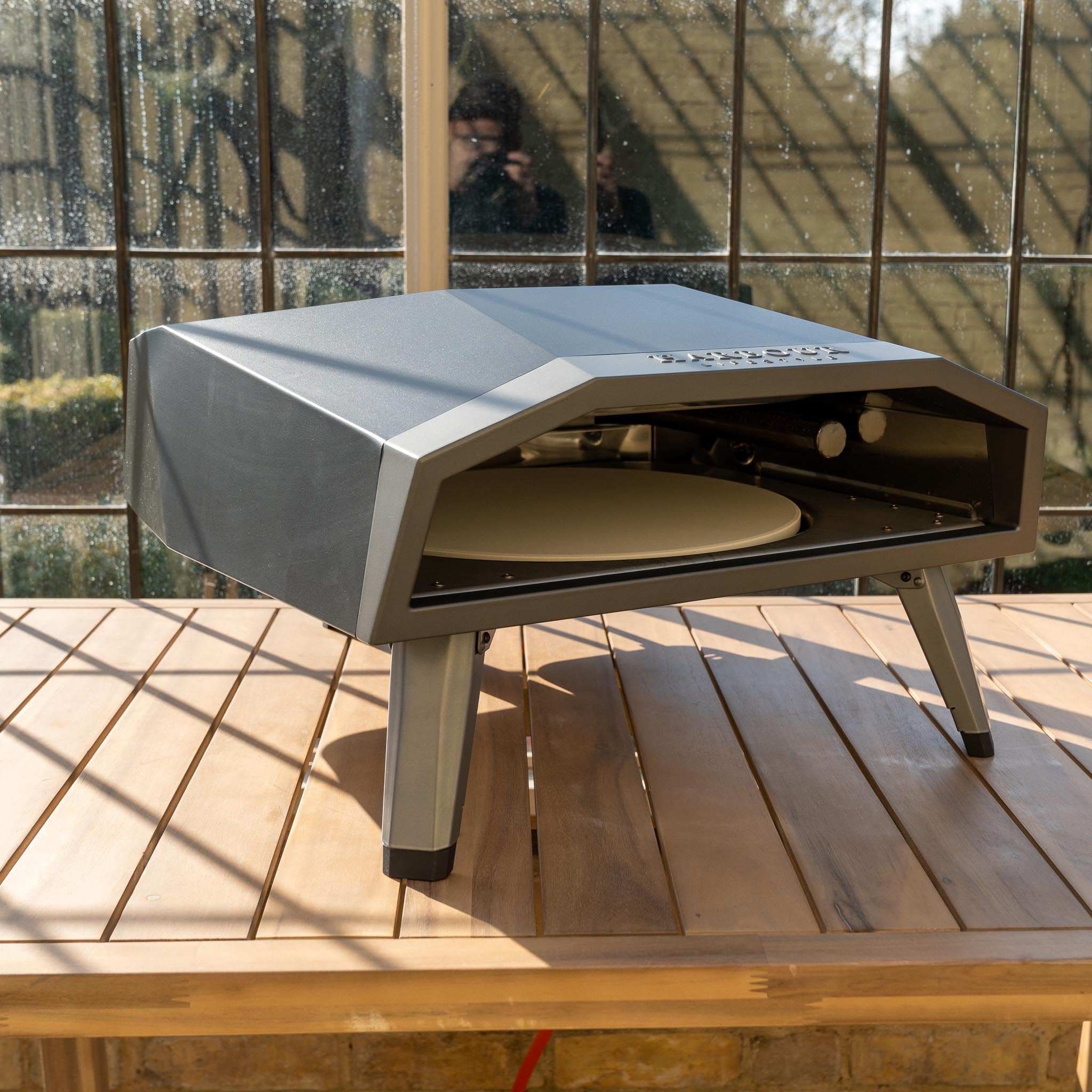 The Juno 16" Pizza Oven Bundle with Stainless Steel Pizza Peel, featuring foldable legs for easy portability, sits on a wooden table in front of a large window.