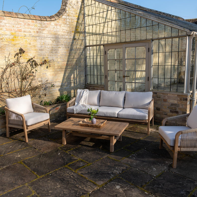 Outdoor patio showcasing the Quay 3 Seat Sofa Set in Linen, featuring wicker furniture and white cushions, complemented by an acacia wood coffee table in front of a brick wall with glass windows.