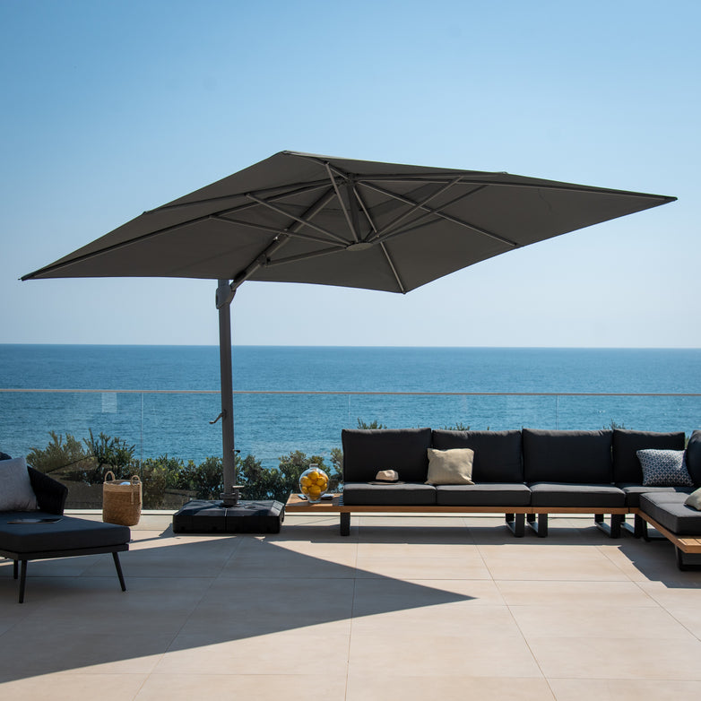 Large outdoor patio with a modern sectional sofa, a Pallas 4m x 3m Rectangular Cantilever Parasol with LED Lighting in Charcoal for garden shade, and an ocean view in the background.