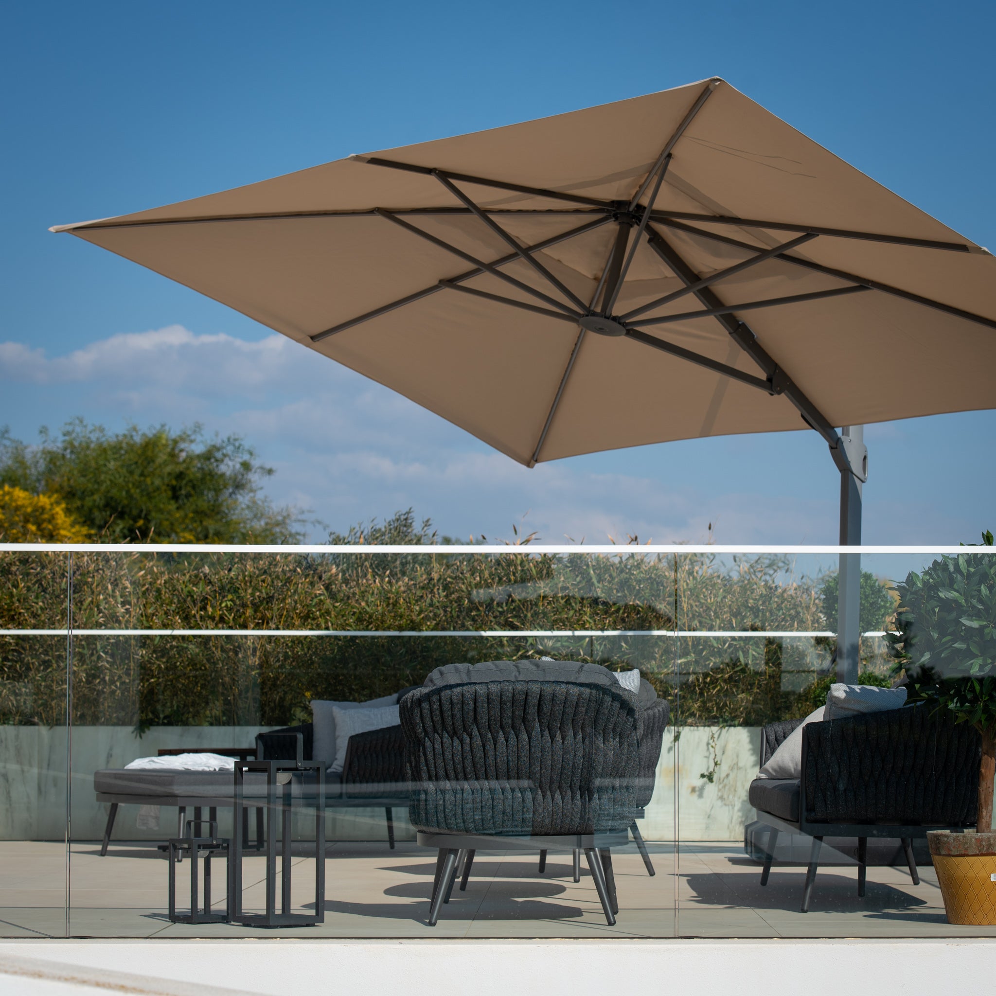 Outdoor patio with wicker furniture and the Pallas 4m x 3m Rectangular Cantilever Parasol with LED Lighting in Beige providing garden shade, glass railing, and greenery in the background.