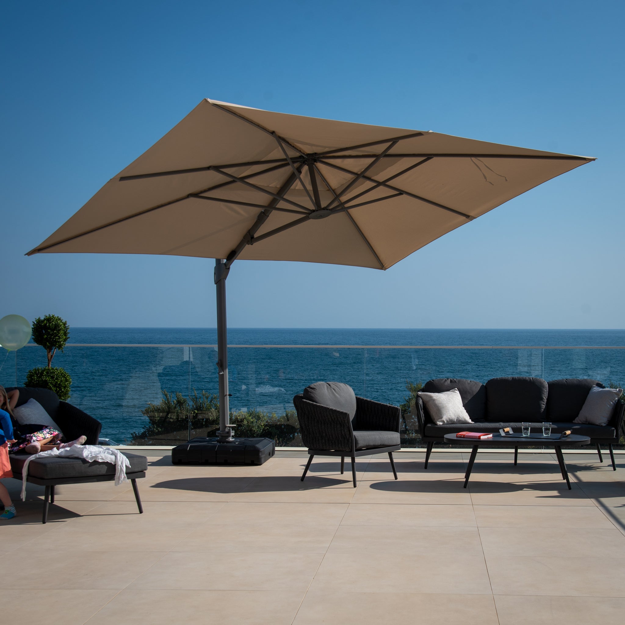 Outdoor seating area with a Pallas 4m x 3m Rectangular Cantilever Parasol with LED Lighting in Beige, black chairs, and a clear view of the ocean under a sunny blue sky.