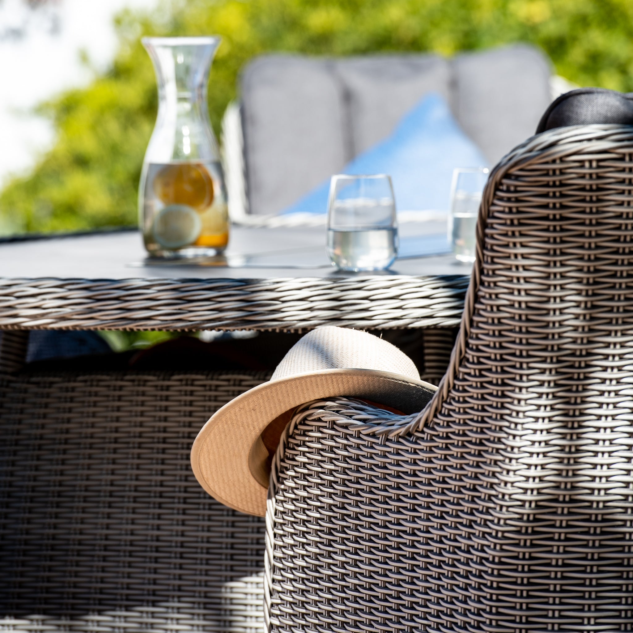 A sun hat hangs on a wicker chair next to the Santiago 6 Seat Oval Rattan Firepit Dining Set in Grey with a pitcher and two glasses outdoors.