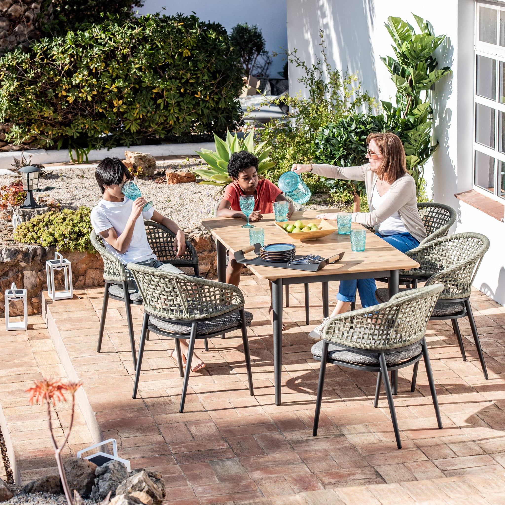 A woman and two children enjoy a meal outdoors at the Kalama 6 Seat Rectangular Dining Set with Teak Table in Olive Green, sitting on comfortable woven chairs, surrounded by greenery and sunlight.