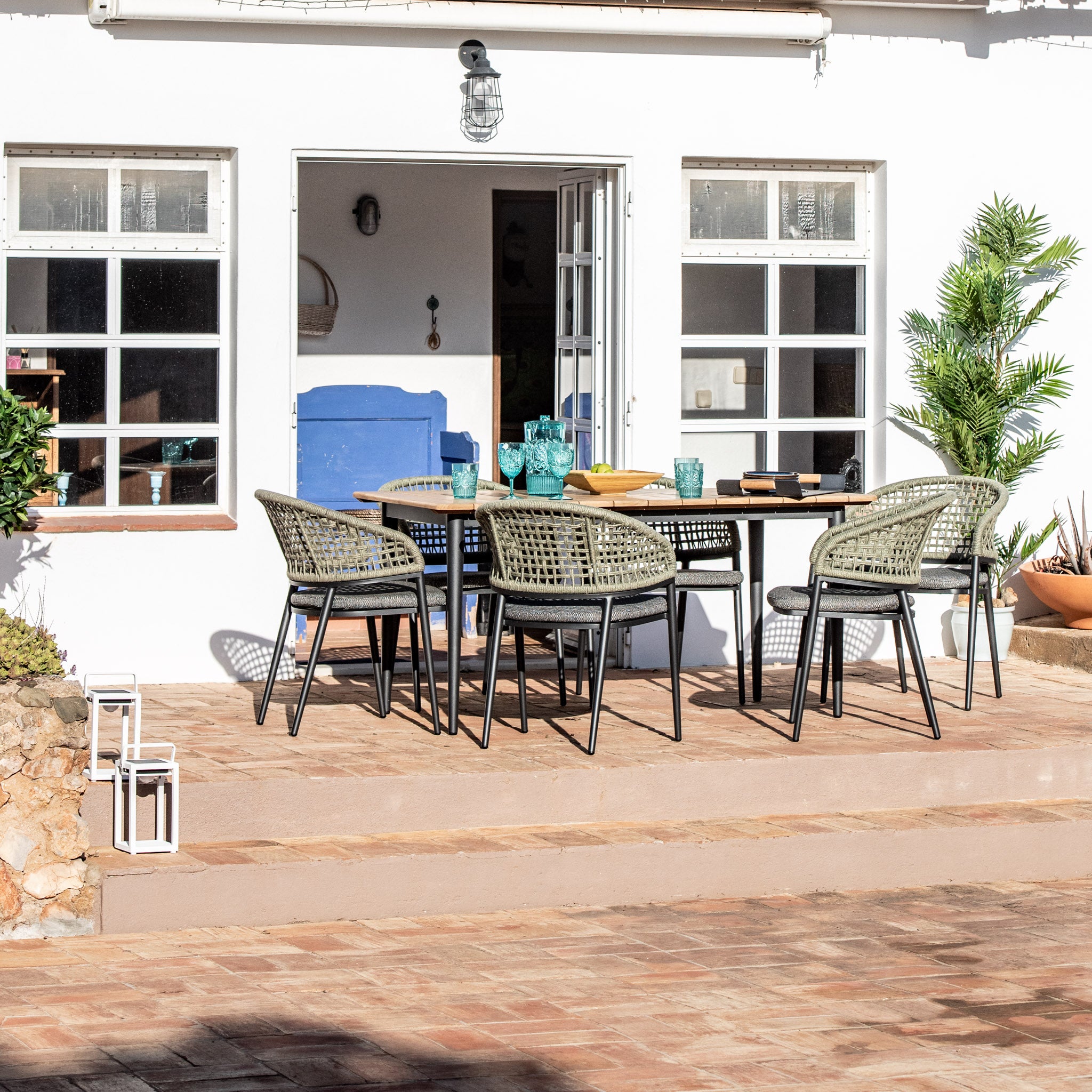 Outdoor dining area featuring the Kalama 6 Seat Rectangular Dining Set with a teak table in olive green, weatherproof aluminum and woven chairs, glassware, white doors, and potted plants around.