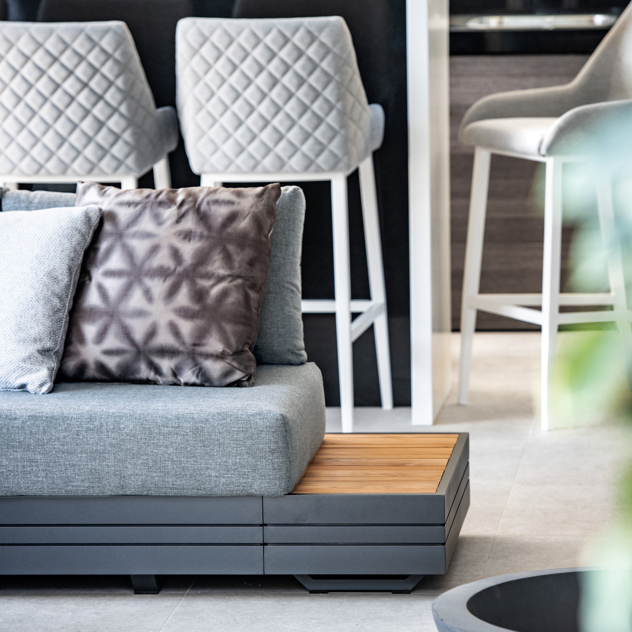 A contemporary living space featuring the Panama Luxury Outdoor Large Corner Group Set in Washed Grey, adorned with patterned cushions and complemented by barstools with quilted backs in the background.