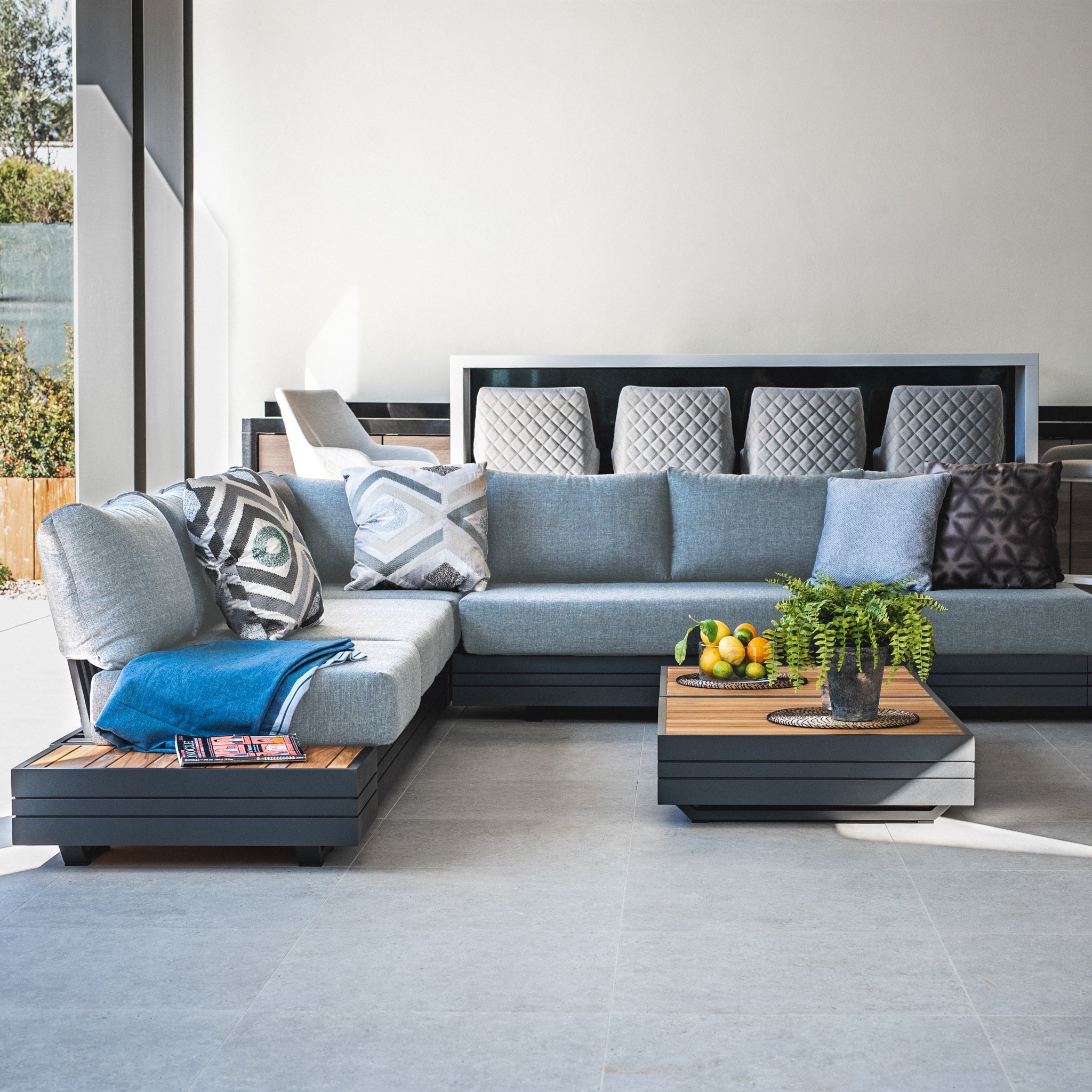The Panama Luxury Outdoor Large Corner Group Set in Washed Grey features a modern modular sofa set with sustainable teak accents, complemented by a plush blue blanket, stylish cushions, a coffee table adorned with fruits, and a refreshing plant.