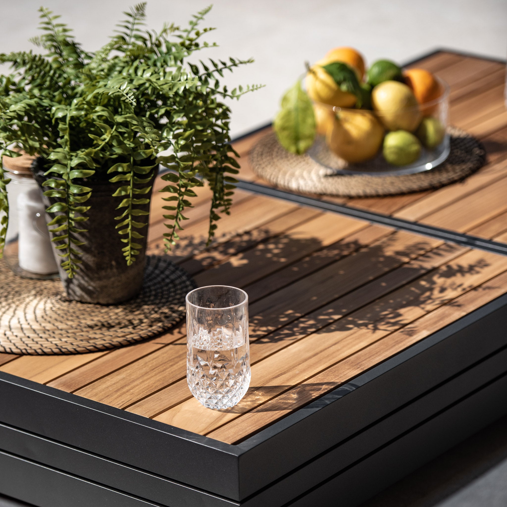 A luxury Panama Teak Coffee Table, adorned with a Teak wooden top, hosts a potted fern, a crystalline glass, and a bowl of assorted fruits.