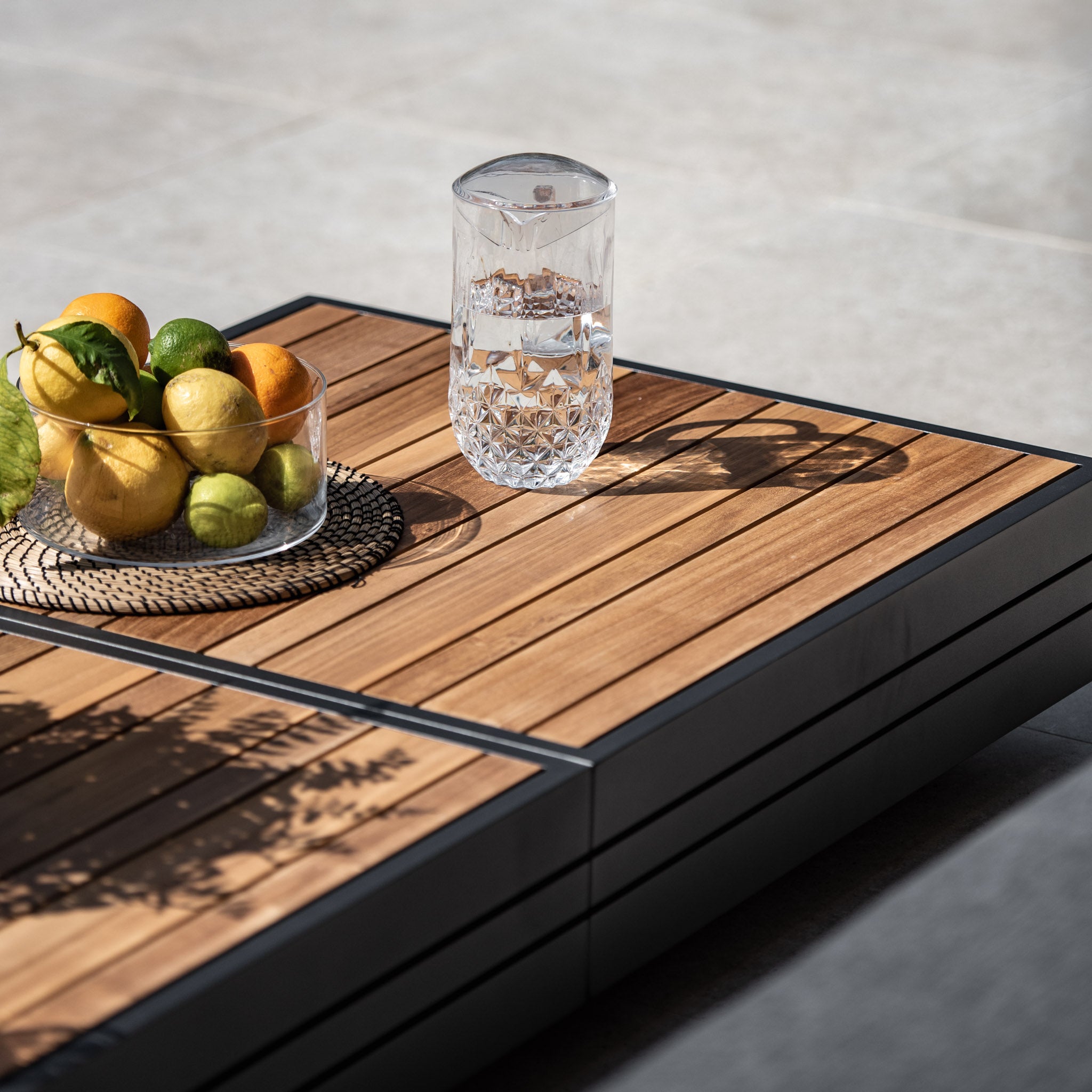 The Panama Teak Coffee Table, adorned with a glass of water and a bowl of mixed citrus fruits featuring lemons and limes, basks in the sunlight, perfectly capturing luxurious outdoor coffee table vibes.
