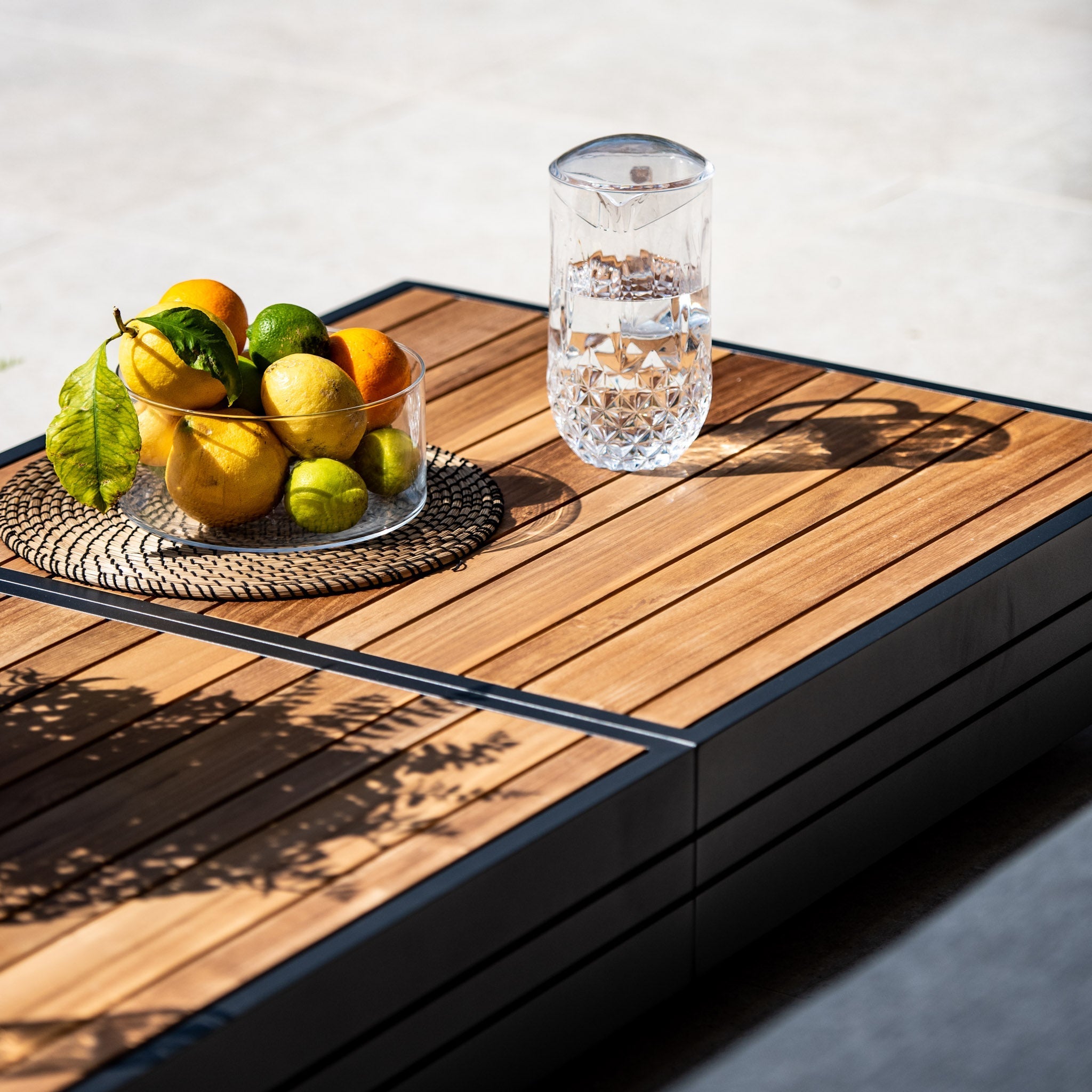 A wooden table with a glass bowl of citrus fruits and a glass of water in a clear, textured glass pitcher sits next to the Panama Luxury Outdoor Large Corner Group Set in Washed Grey on a sunny day.