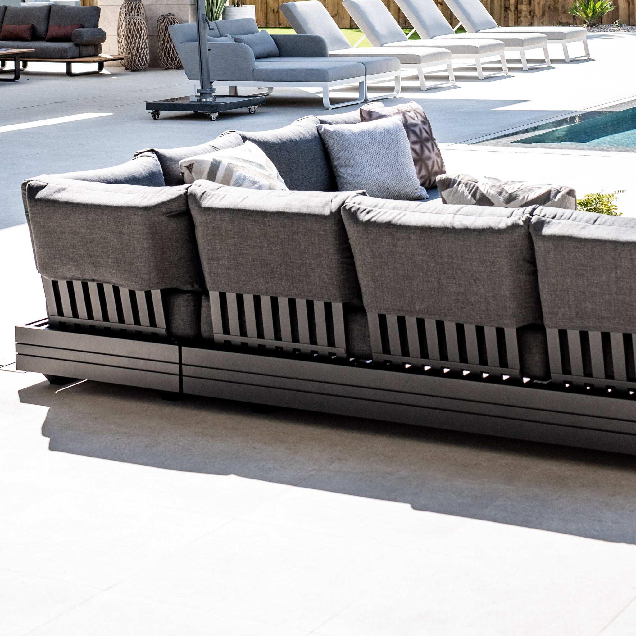 Outdoor seating area with the Panama Luxury Outdoor Large Corner Group Set in Charcoal by a pool, surrounded by lounge chairs and sunlight.