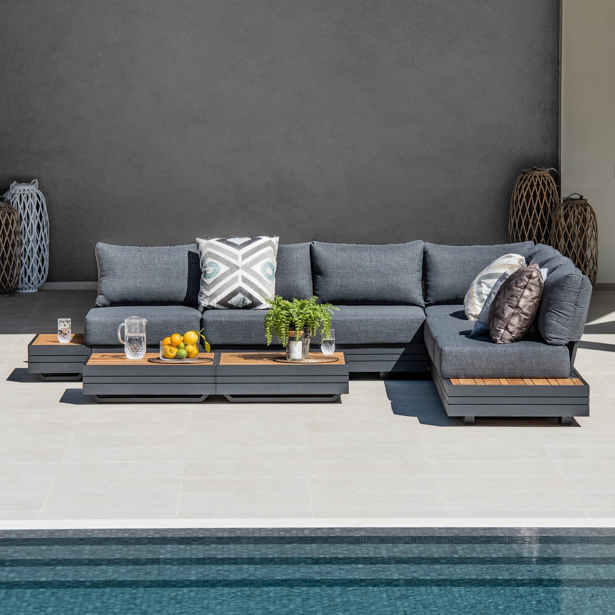 Modern patio with a luxury outdoor furniture setup featuring the Panama Luxury Outdoor Large Corner Group Set in Charcoal, accented with geometric and textured pillows. Sustainable teak wooden coffee tables add warmth to the space, while a serene pool shimmers in the foreground.