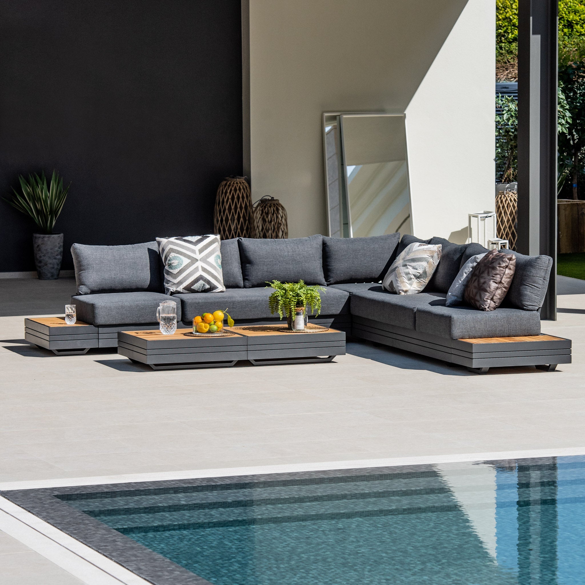 Modern outdoor seating area featuring the Panama Luxury Outdoor Large Corner Group Set in Charcoal, accompanied by a sustainable teak coffee table adorned with fruits and a plant, situated near a pool and mirror.
