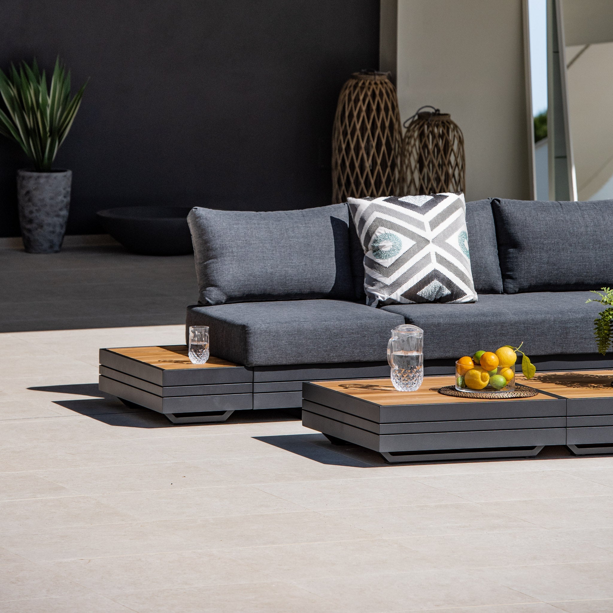 Modern outdoor seating with dark cushions, a geometric pillow, two glasses of water on tables, and a tray of fruit, all beautifully arranged on a Panama Luxury Outdoor Large Corner Group Set in Charcoal.