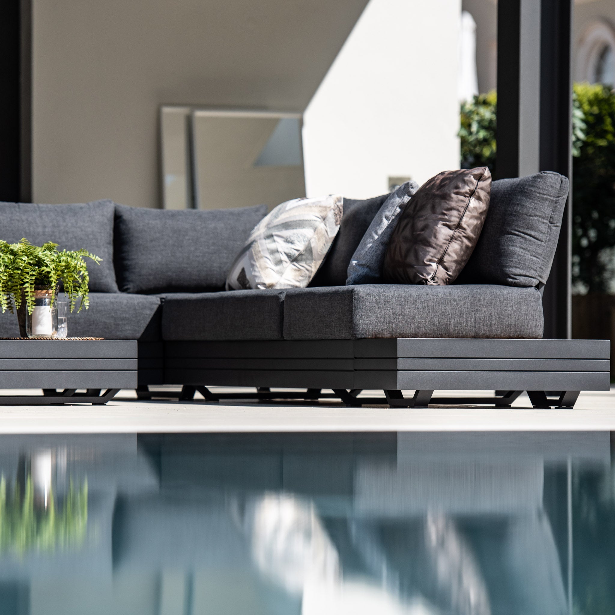 The Panama Luxury Outdoor Large Corner Group Set in Charcoal includes a modular sofa set made from sustainable teak, featuring dark cushions next to the pool, decorated with a plant and pillows.