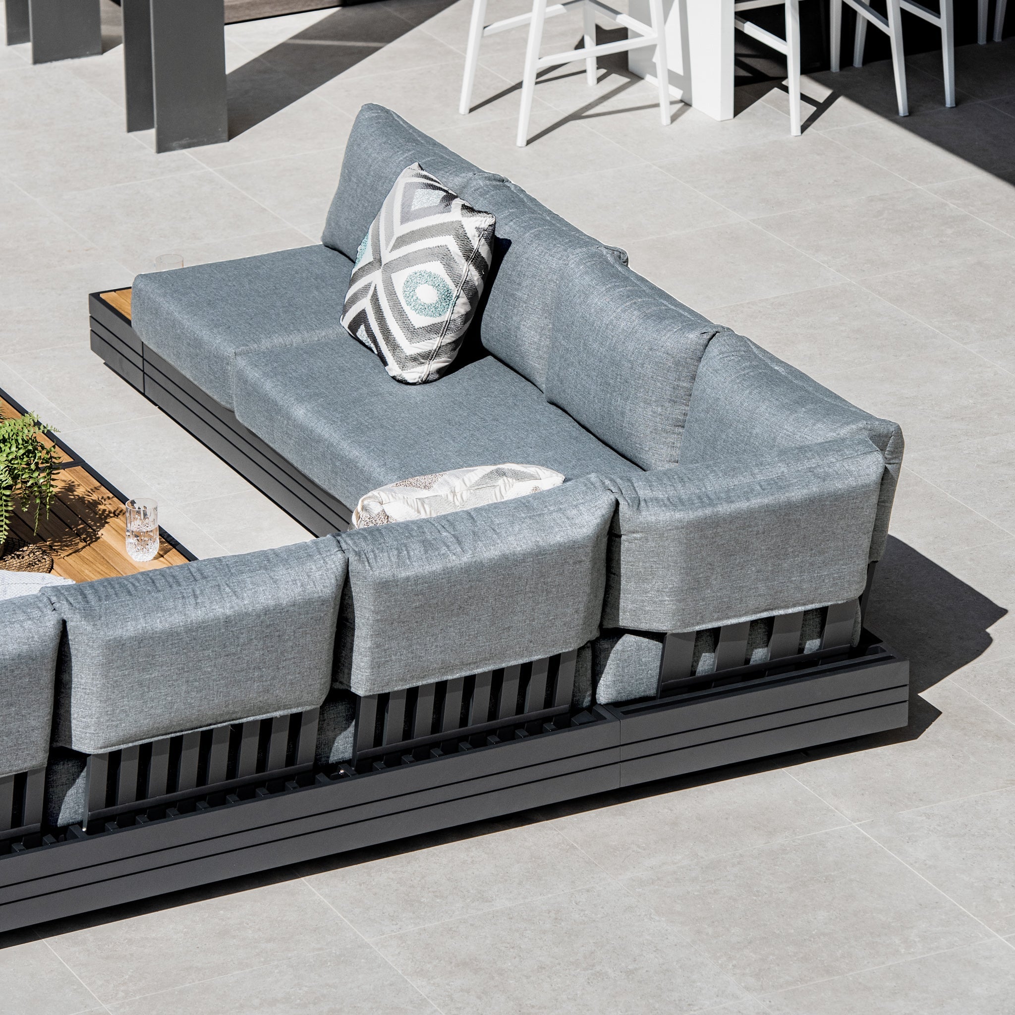 The Panama Luxury Outdoor Large Corner Group Set in Washed Grey, featuring gray cushions and patterned pillows, surrounds a sustainable teak coffee table on a tiled patio, creating an inviting and eco-friendly space.