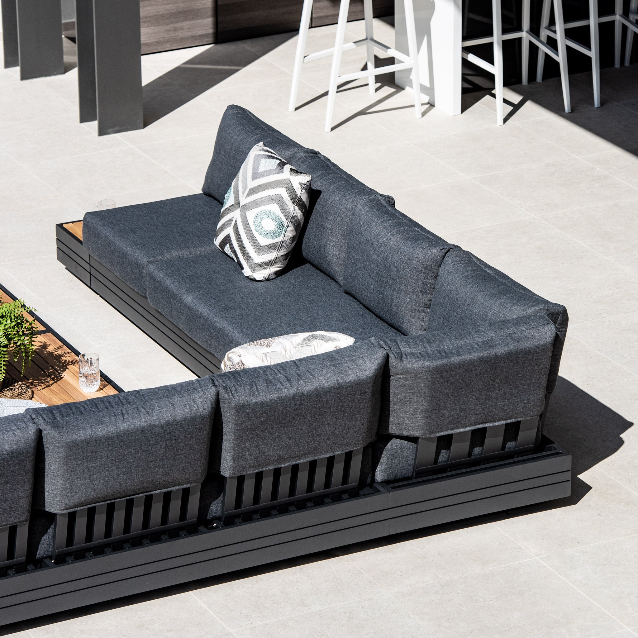 The Panama Luxury Outdoor Large Corner Group Set in Charcoal is a luxury outdoor furniture piece that features modern modular design with gray cushions, a patterned throw pillow, and a wooden coffee table adorned with plants crafted from sustainable teak.