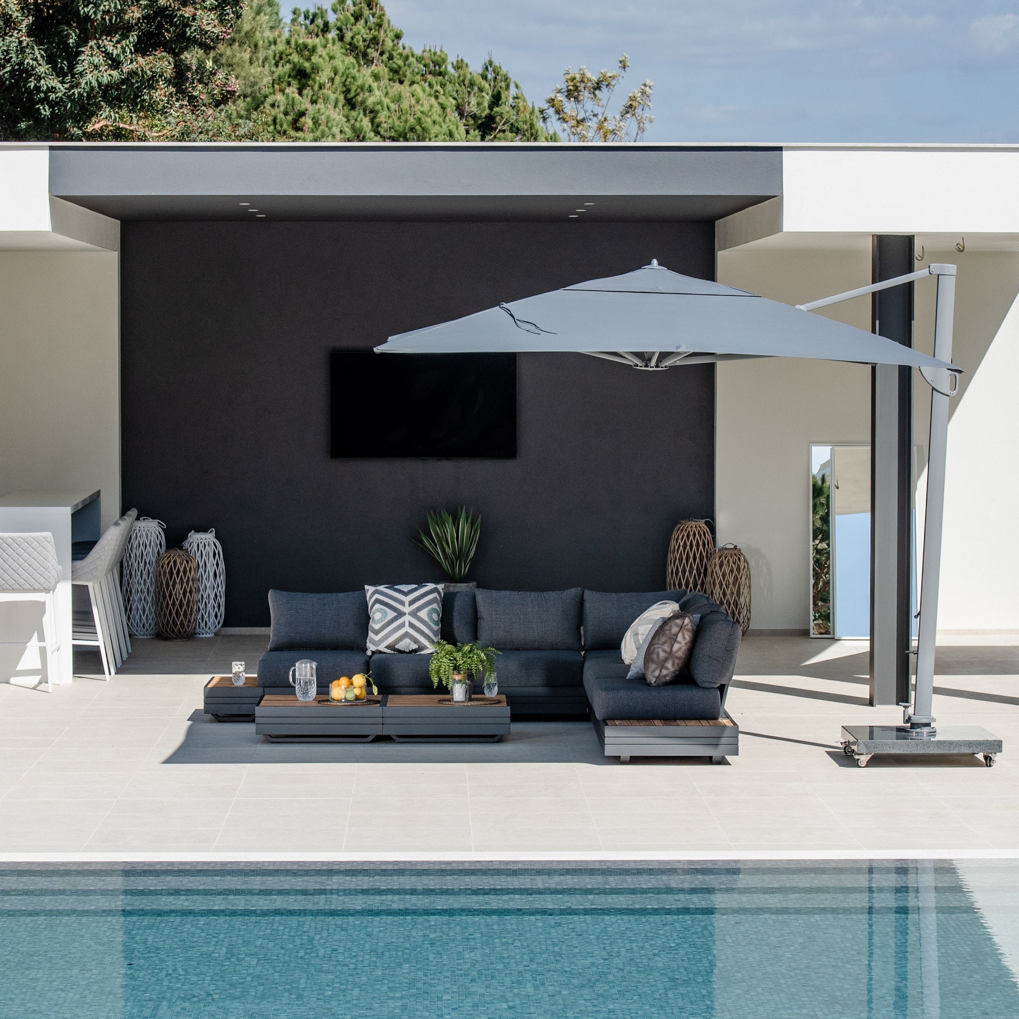Modern outdoor patio with a pool, the Panama Luxury Outdoor Large Corner Group Set in Charcoal, and a large umbrella featuring luxury outdoor furniture made from sustainable teak, surrounded by green plants under a clear sky.