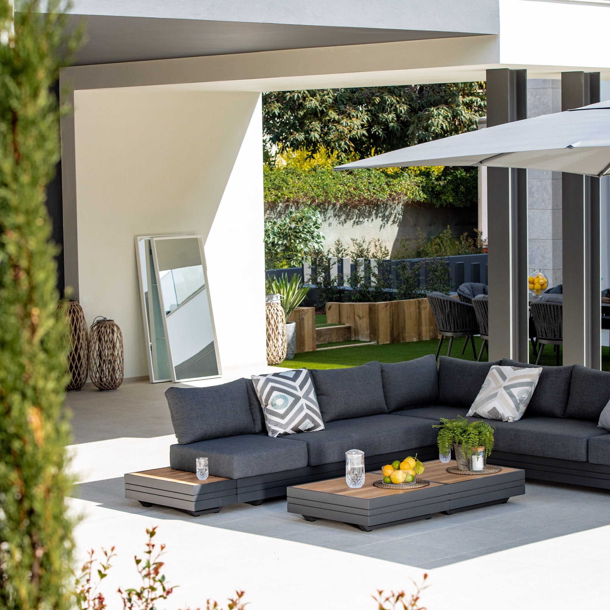 Modern patio featuring the Panama Luxury Outdoor Large Corner Group Set in Charcoal, a low table, potted plants, a large mirror, and an umbrella in a sunny backyard. Enjoy luxury outdoor furniture that combines style and comfort for the perfect alfresco retreat.