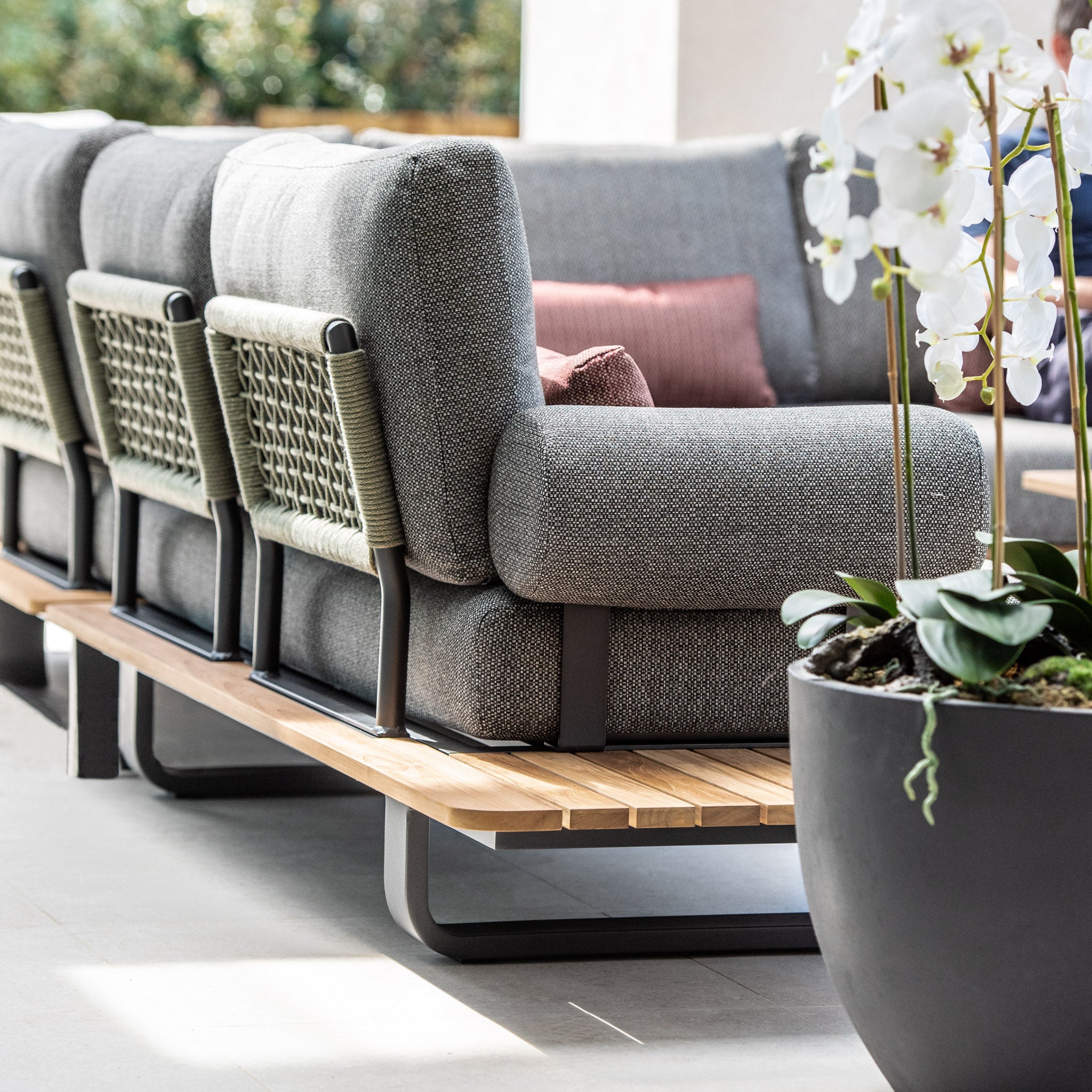 Modern outdoor seating featuring a Bay Large Corner Group Set with 4 waist pillows and a teak table in olive green, complemented by grey cushions, a wooden base, and a potted orchid plant in the foreground.