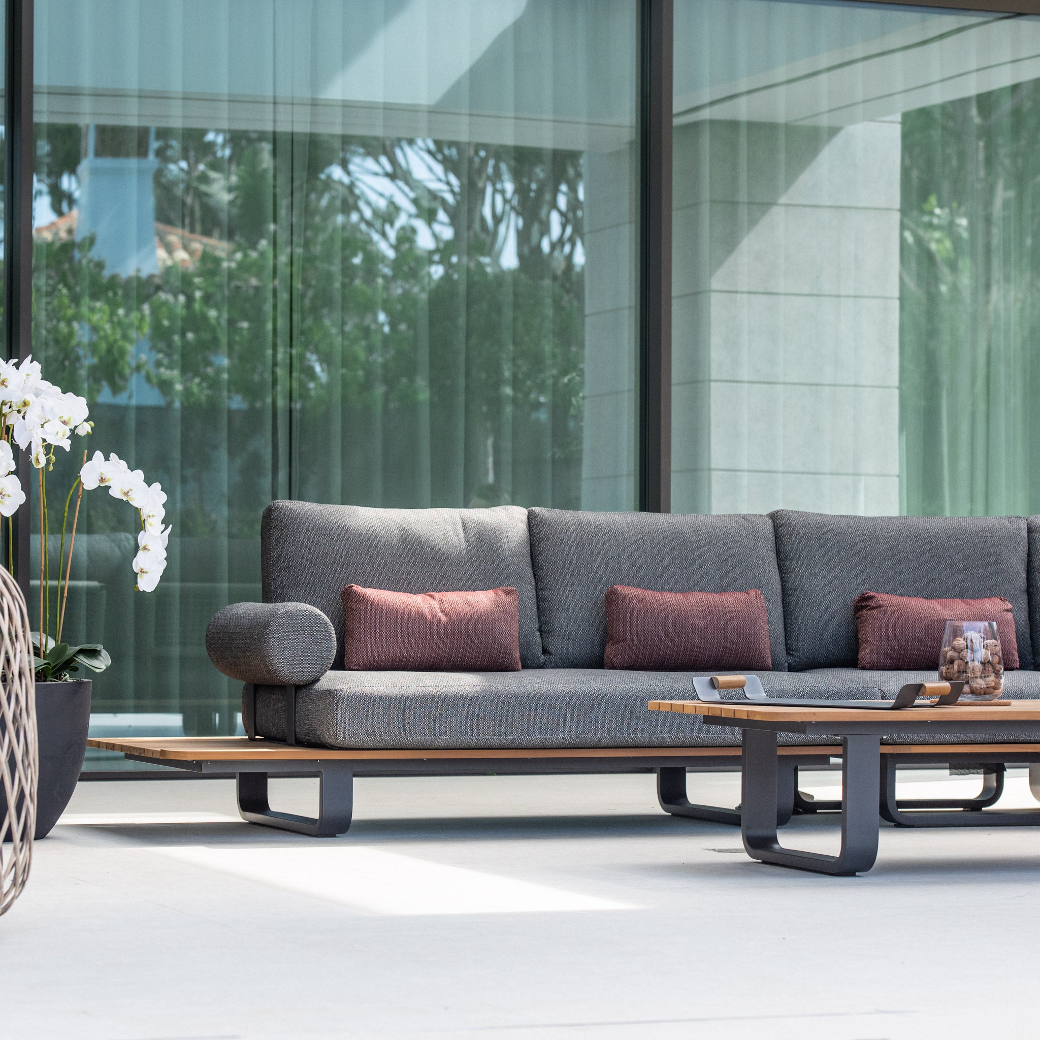 Modern outdoor seating area with a Bay Large Corner Group Set in olive green, featuring four waist pillows, a teak table, and potted orchids in a sleek setting.