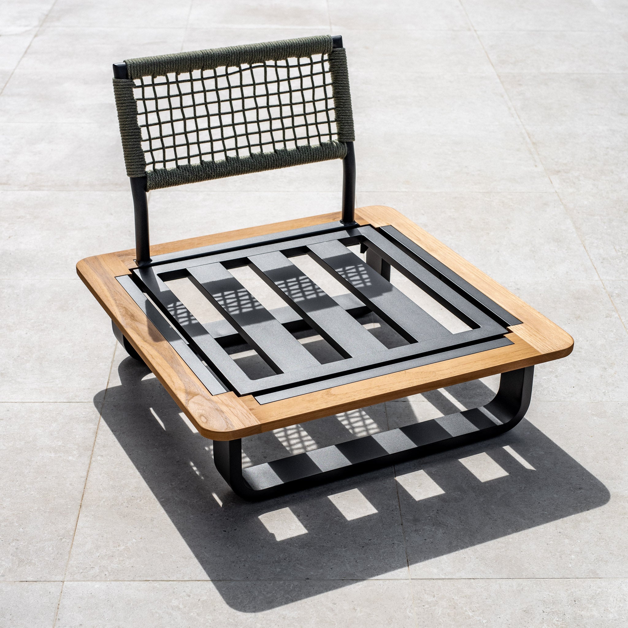 A contemporary, low-profile Bay Large Corner Group Set with a wooden frame, black metal strips, and a woven backrest paired with a sleek teak table in olive green on a tiled floor, complete with 4 waist pillows.