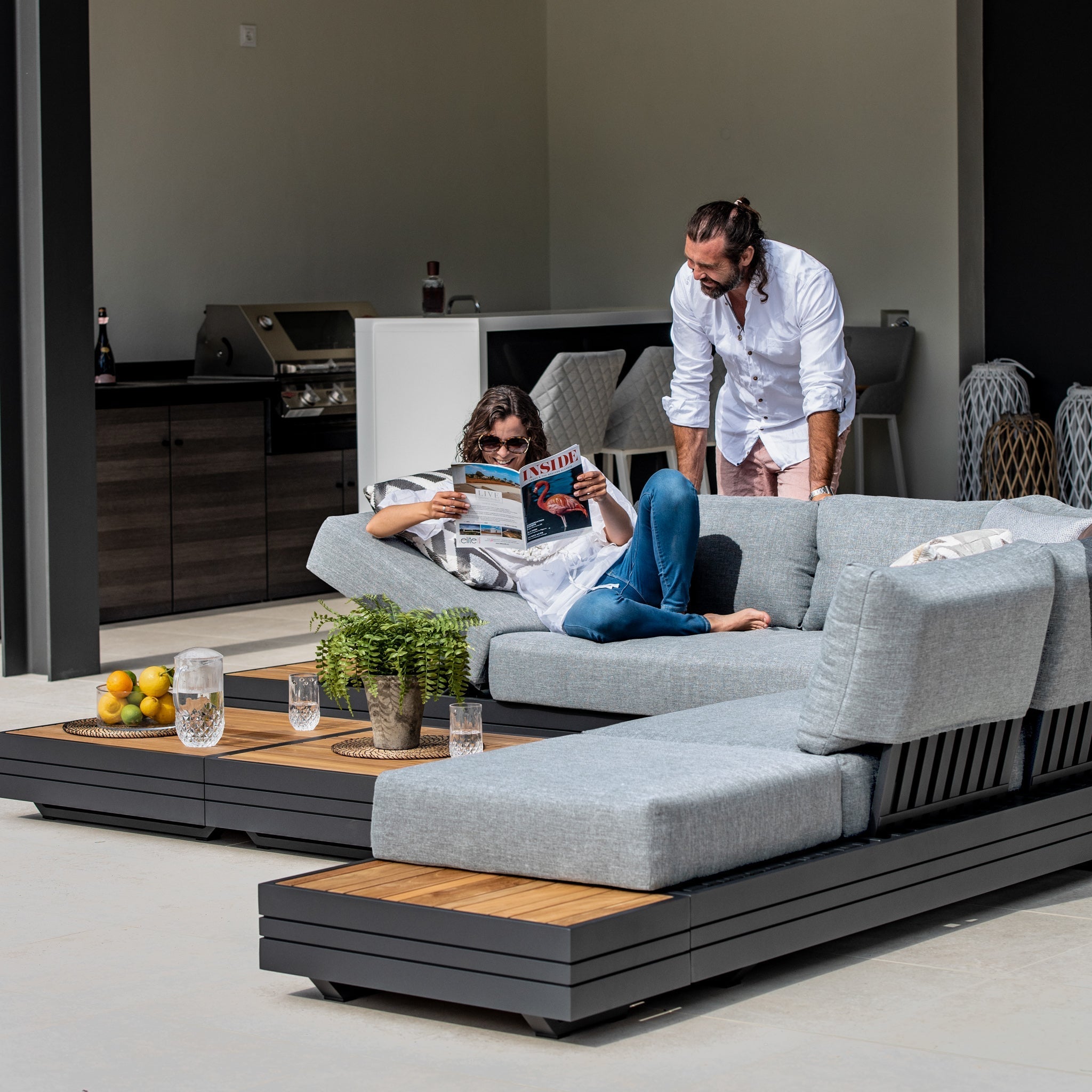Two people relax on a Panama Luxury Outdoor Large Corner Group Set in Washed Grey; one reads a magazine while the other stands nearby, engaging in conversation. The stylish ensemble, crafted from sustainable teak, enhances the tranquil ambiance.