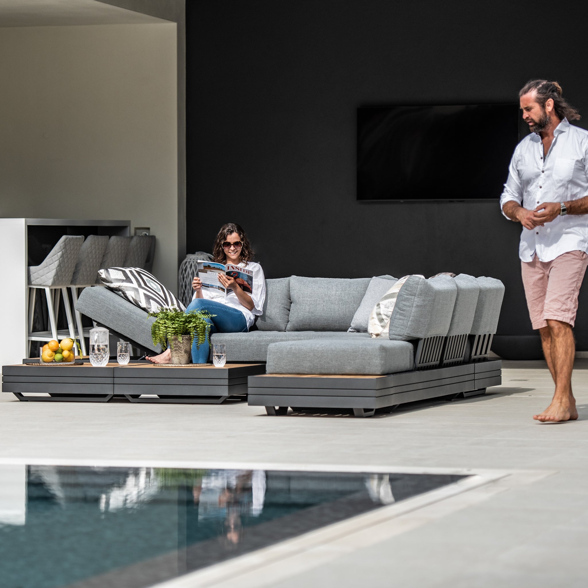 A woman reads on a modern patio couch from the Panama Luxury Outdoor Large Corner Group Set in Washed Grey near a pool, while a man stands nearby in the stylish setting. The modular sofa set features sustainable teak accents, seamlessly blending elegance and eco-friendliness.