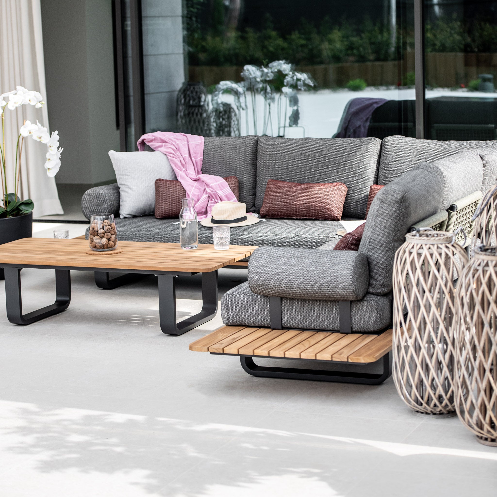 Modern outdoor patio with a Bay Corner Group Set, featuring 4 waist pillows and a teak coffee table in olive green, complemented by decorative pillows made of olive green acrylic rope and wicker lanterns.